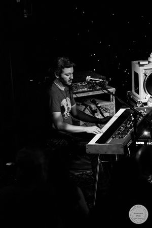 Black and white photo of male musician playing keyboard synthesizer on stage during a live performance.