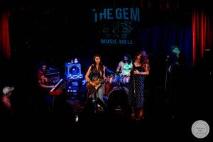 Live band performance at The Gem Music Hall with musicians on stage under blue and red stage lighting.