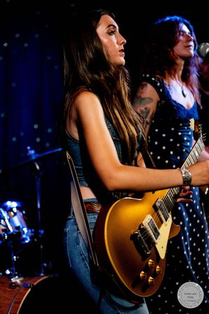Young female guitarist plays gold Les Paul electric guitar on stage under warm concert lighting.