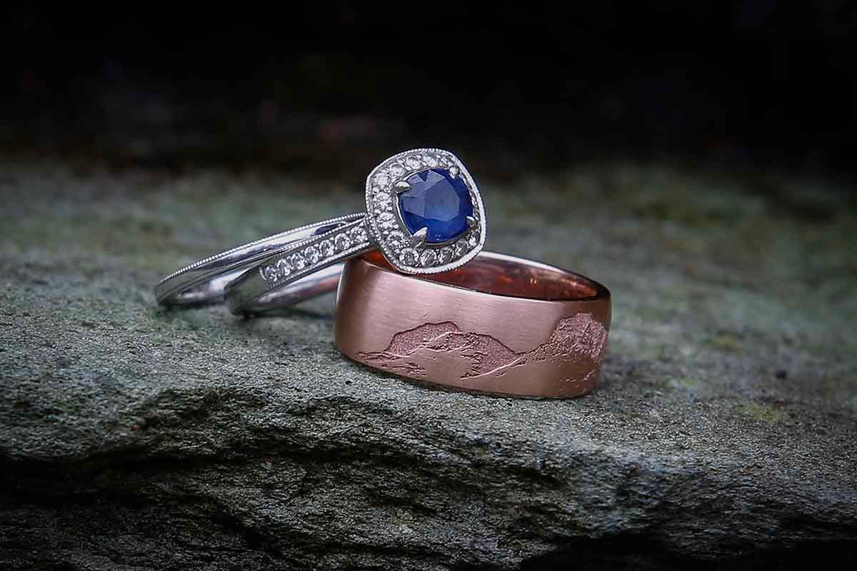 Two elegant rings on stone: a silver halo sapphire engagement ring and a rose gold mountain landscape wedding band depicting the Langdales