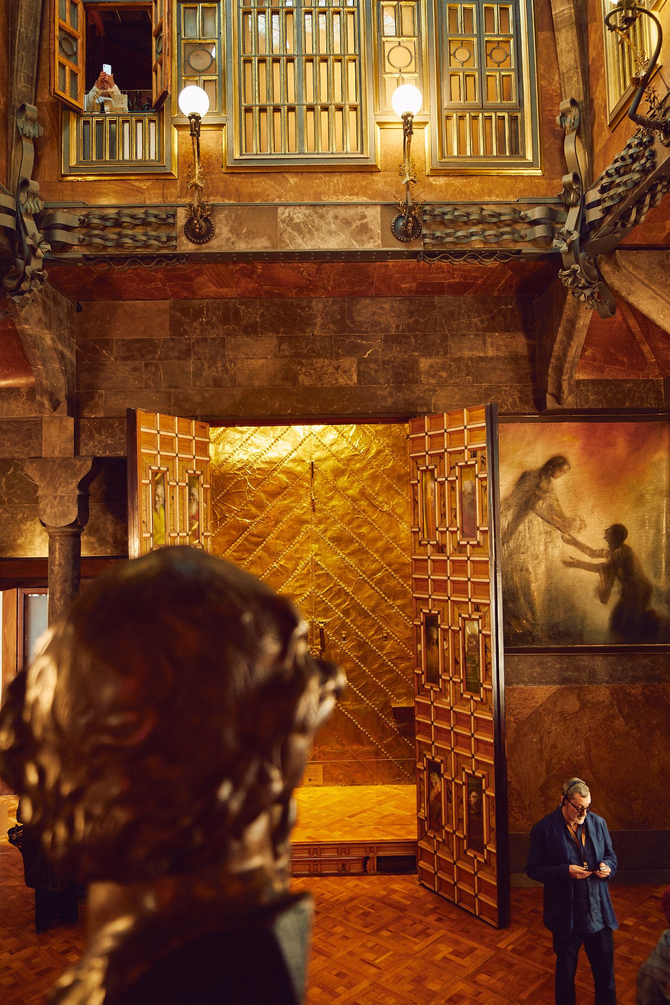 Ornate golden doors open in a grand historic interior with warm lighting, artwork, and visitors exploring the space.