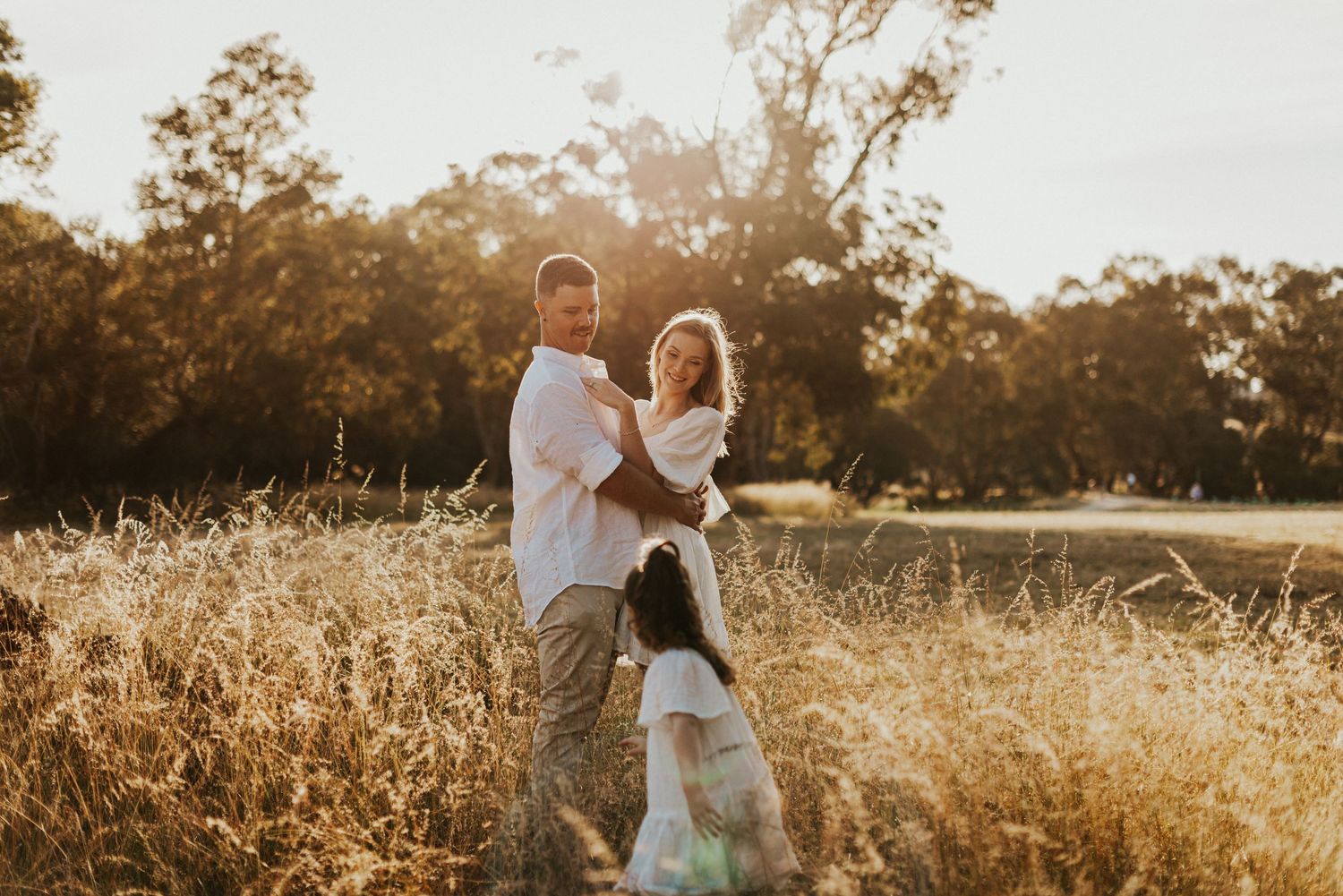 Family posing in golden field at sunset, parents holding baby while young child stands nearby.