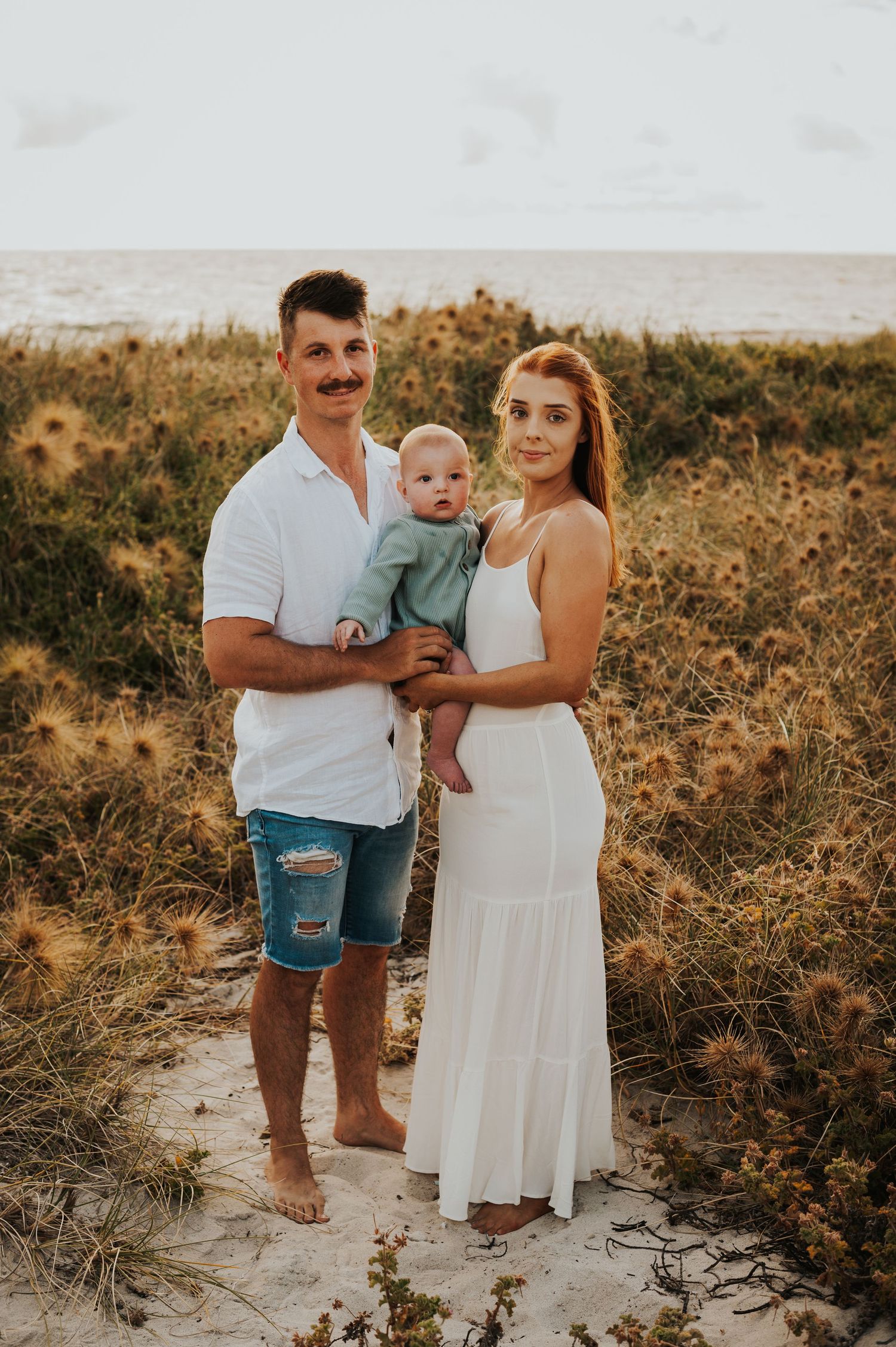 Young family with baby posing on sandy beach path surrounded by coastal shrubs at sunset.