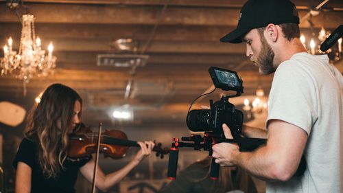 A cameraman films a young woman playing violin in a rustic venue with elegant chandeliers overhead.