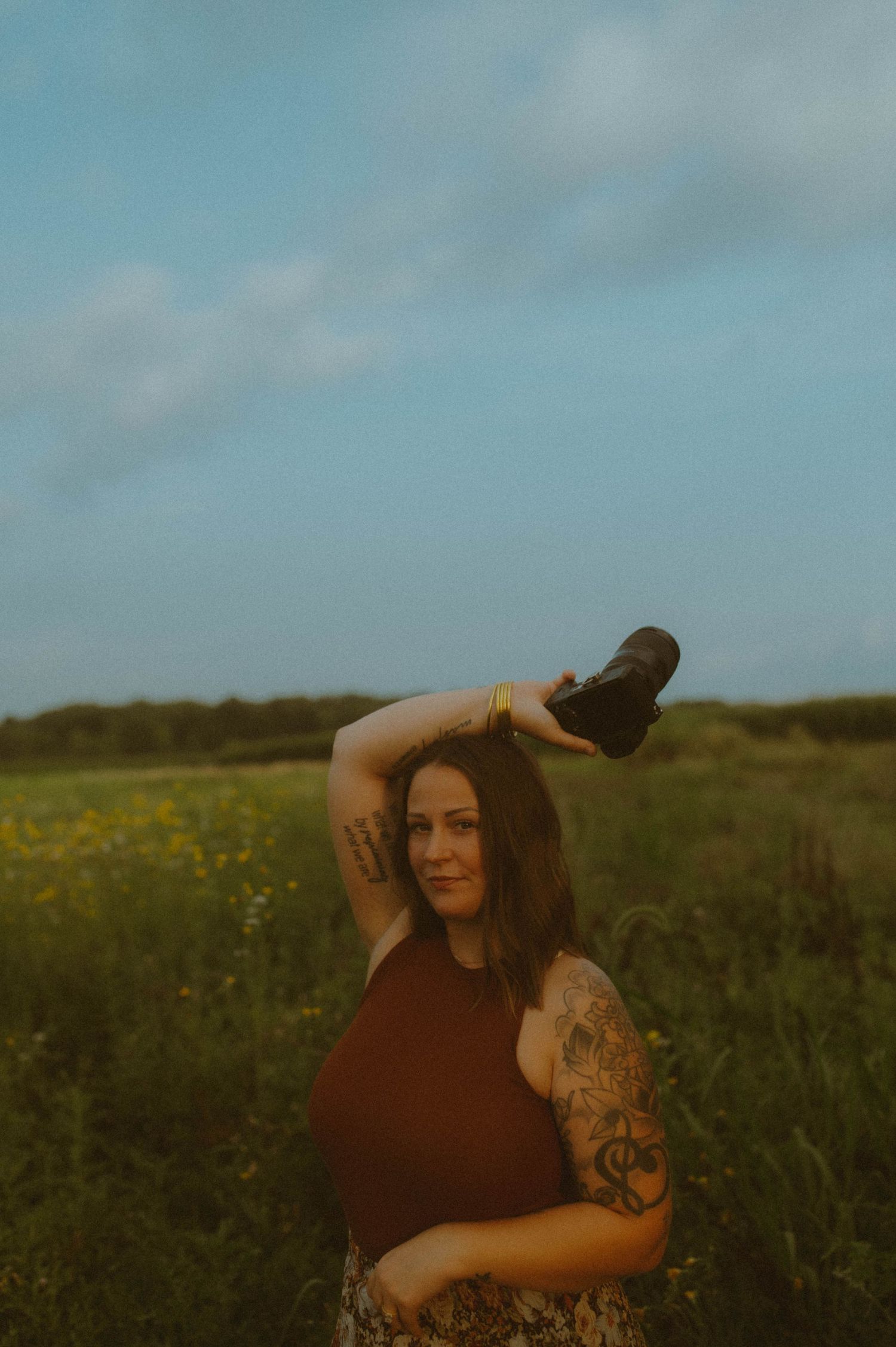 A tattooed woman in a brown top holds a camera in a wildflower field at dusk.