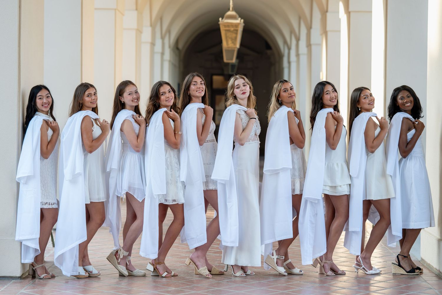 Group of eleven women in matching white dresses posing in an elegant arched corridor with chandelier.