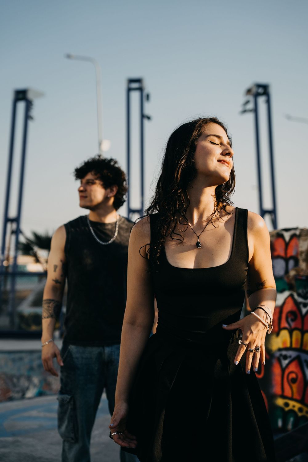 A tattooed couple in black outfits pose outdoors near colorful graffiti wall under a clear sky.
