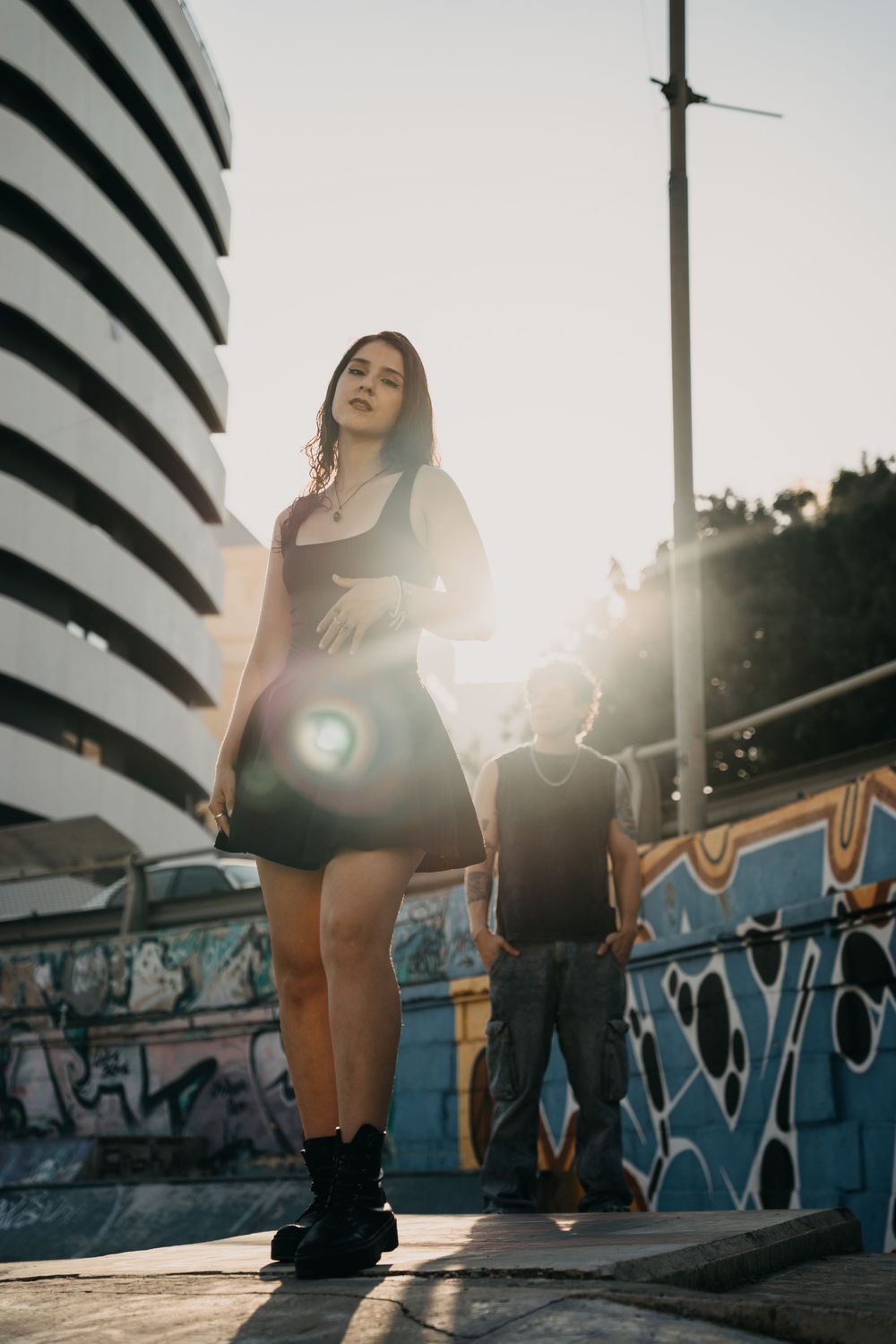 Young woman in black dress and boots poses on urban street with sunflare and graffiti wall backdrop.