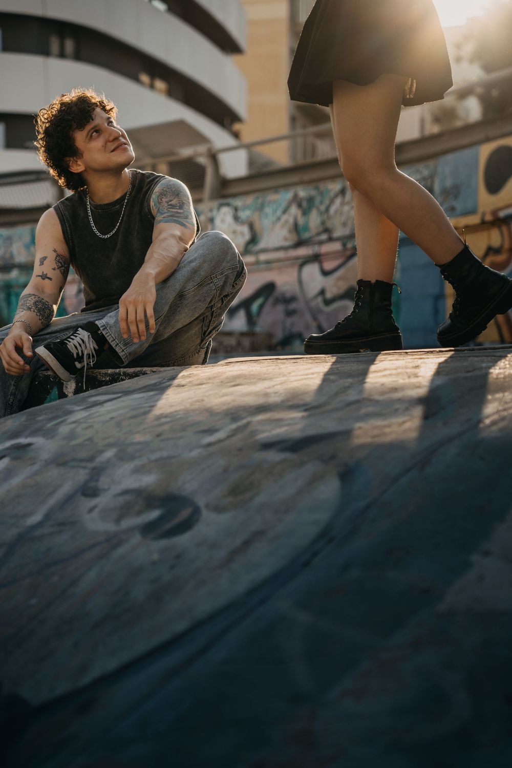 Tattooed skater guy sitting on a ramp holding a skateboard, looking up at a person standing nearby at an urban skate park.
