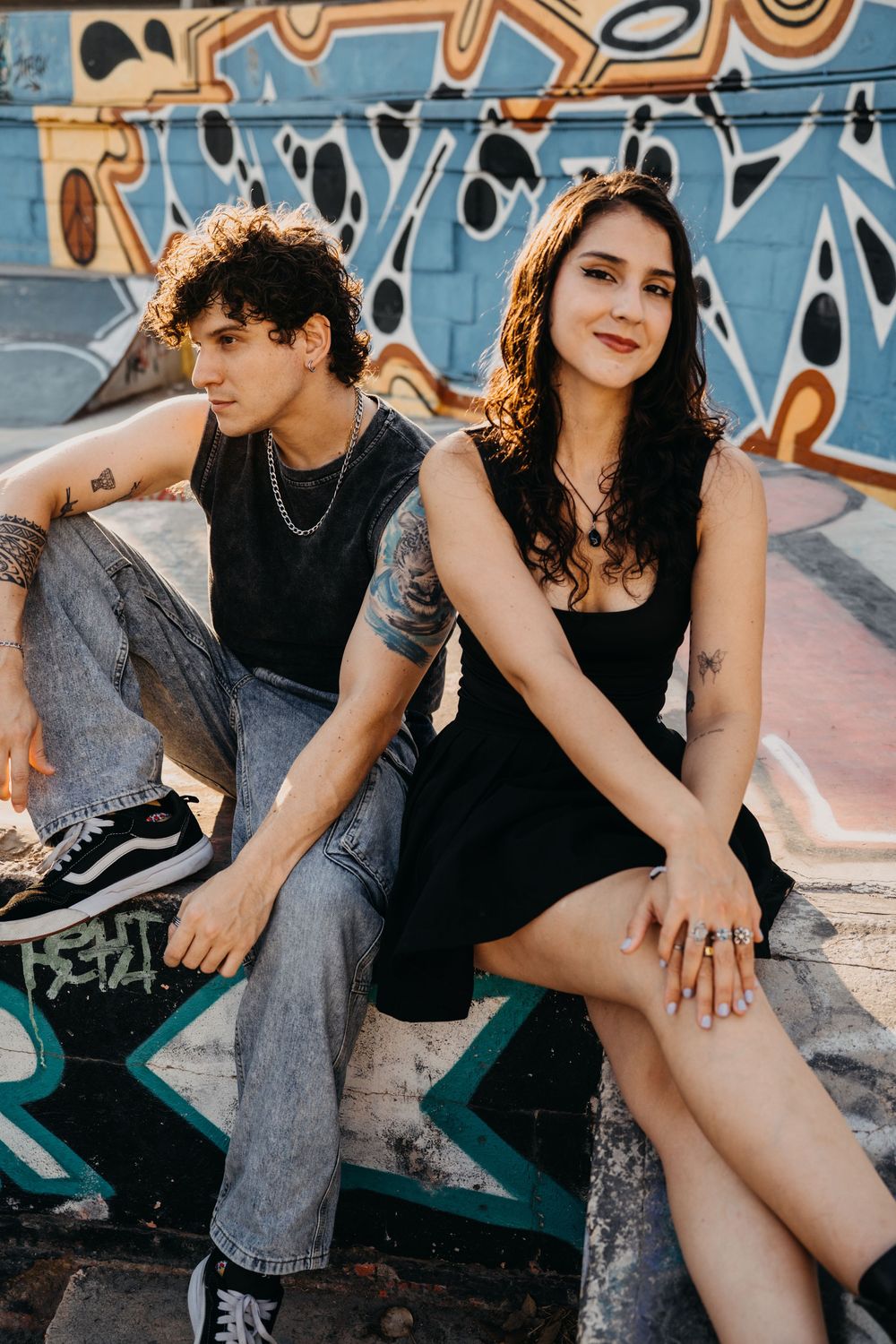 Two young people in black outfits sit against a colorful graffiti wall at a skate park.