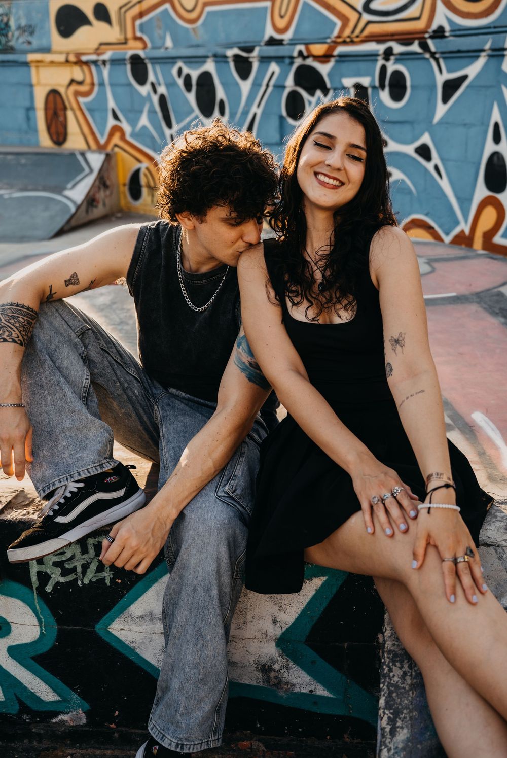 A smiling couple with tattoos sitting at a graffiti-covered skate park, wearing casual black outfits.