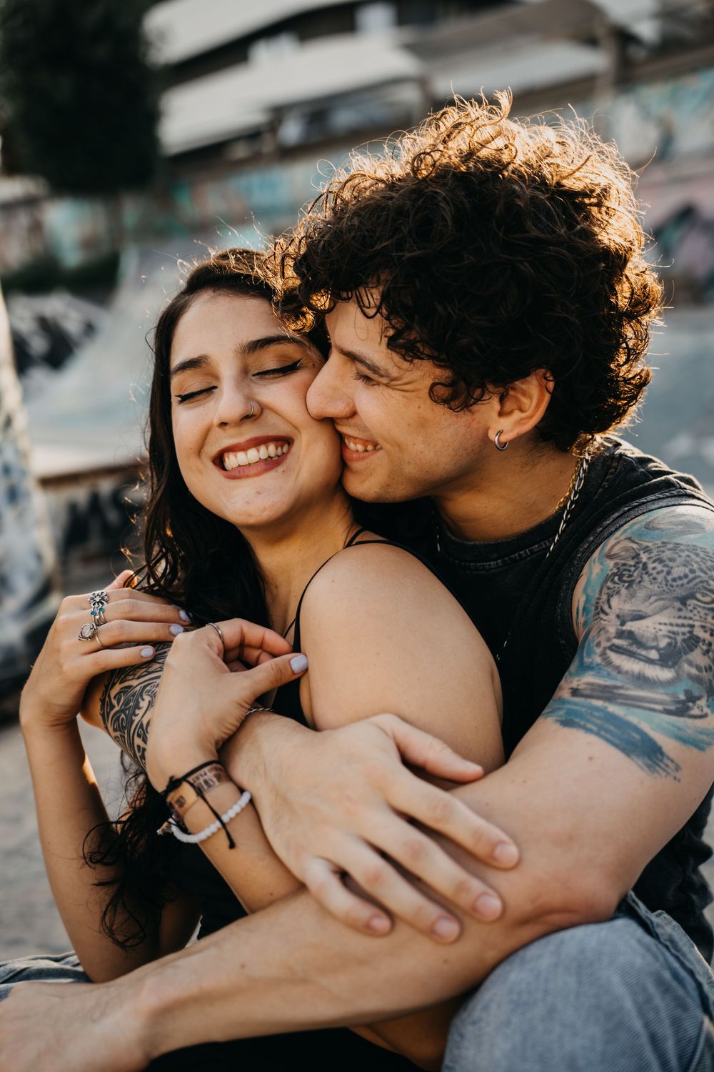 A tattooed couple embracing and smiling outdoors, the man kissing the woman's cheek affectionately.