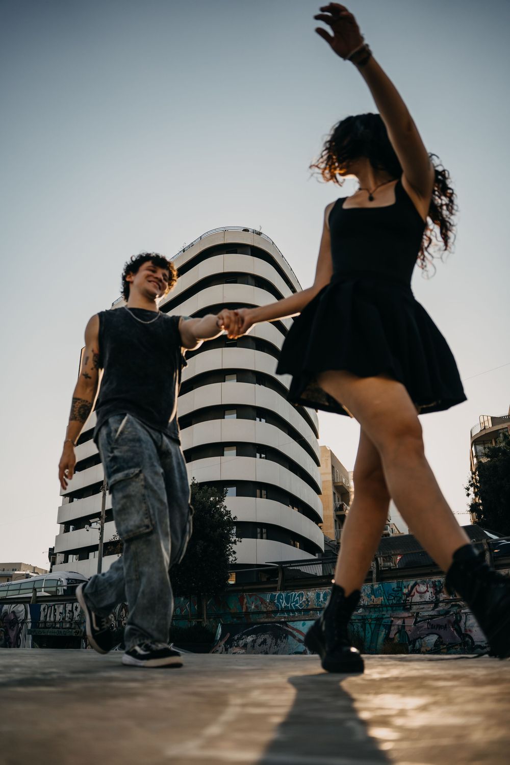 Two young people dancing outdoors near urban graffiti walls, woman in black dress twirling, man in jeans smiling at sunset.