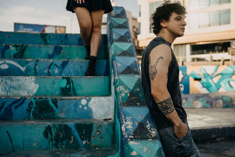 Young tattooed man in sleeveless shirt stands by colorful graffiti-covered blue stairs in urban setting.