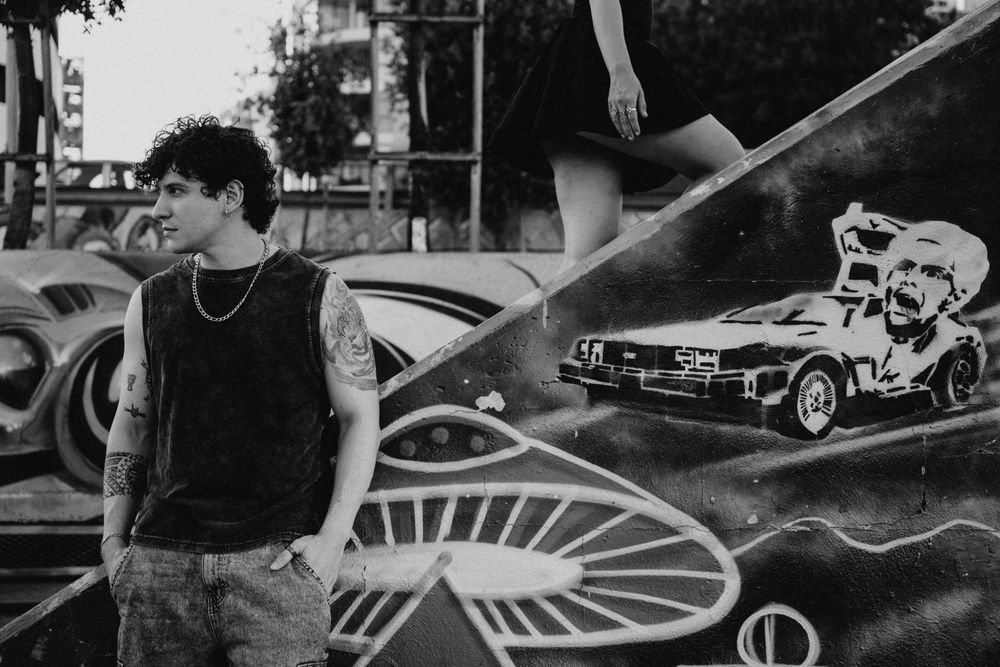 Young man with tattoos poses before a graffiti mural featuring a DeLorean car in black and white photo.