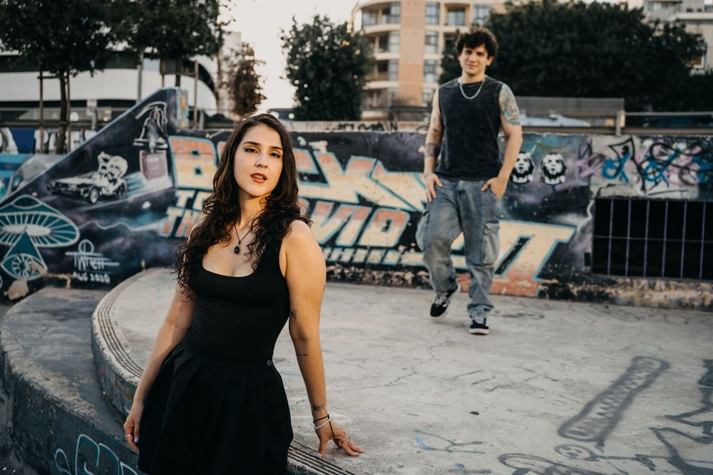 Two young adults pose at a graffiti-covered skatepark, woman in black dress foreground, tattooed man standing behind.