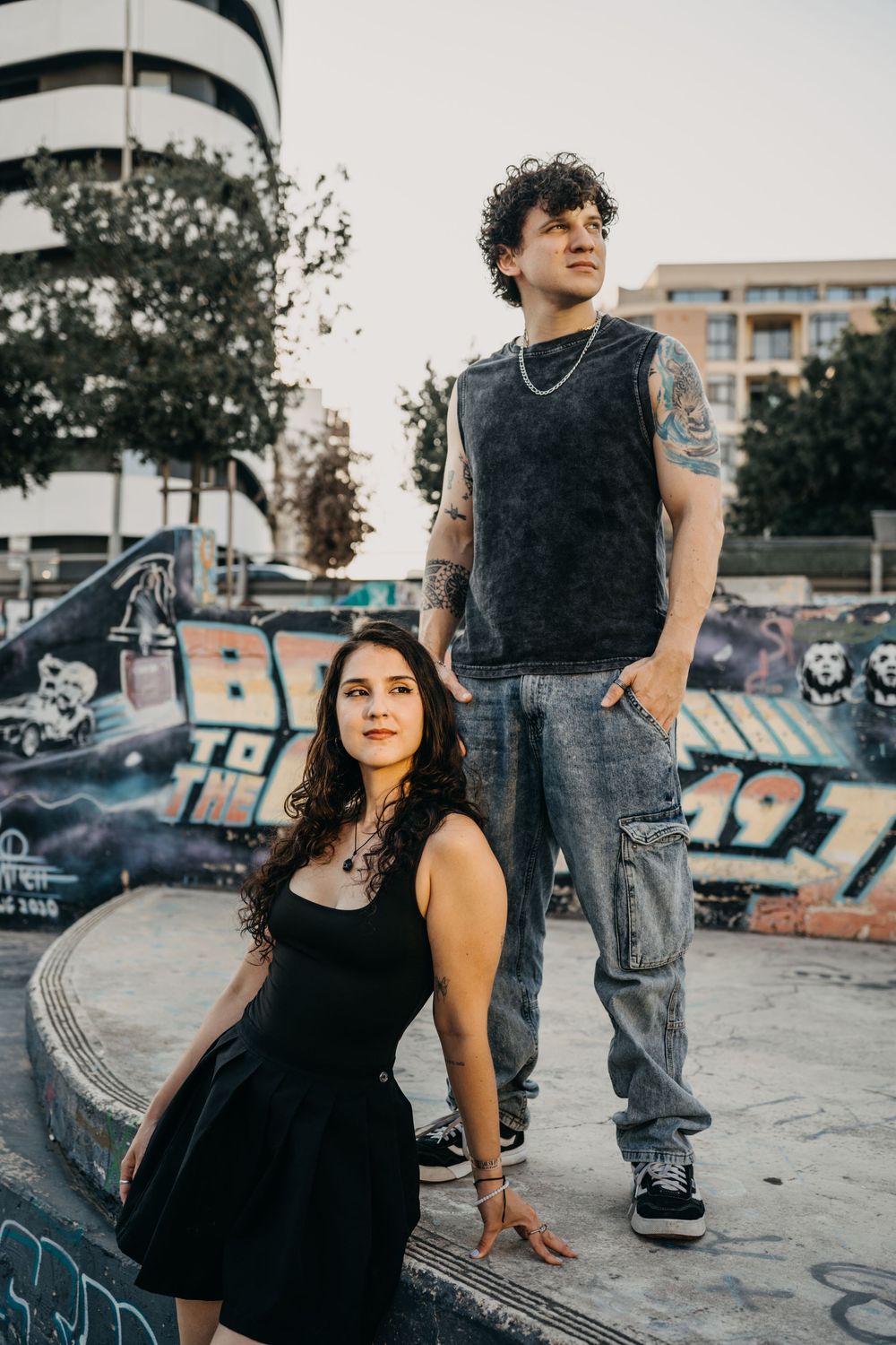 A tattooed man and woman pose at a graffiti skate park, she sits in black dress, he stands in jeans and sleeveless top.