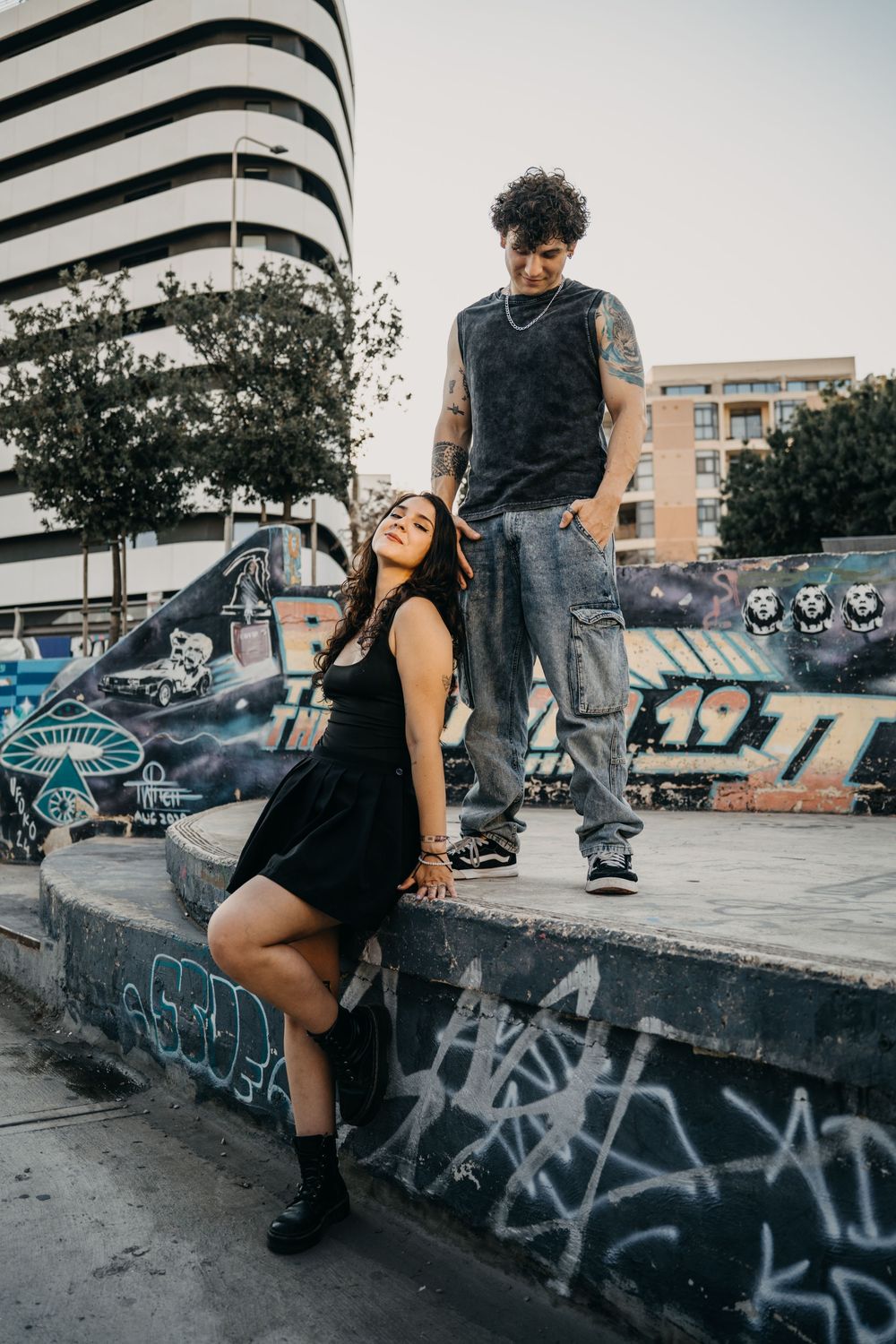 A young couple poses at a graffiti-covered skate park, woman in black dress seated, tattooed man standing above her.