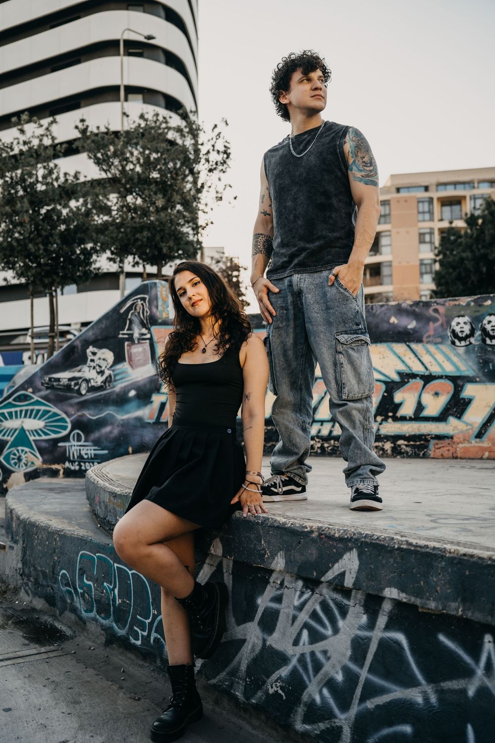 A tattooed couple poses at a graffiti-covered skate park, woman in black dress, man in jeans and sleeveless shirt.