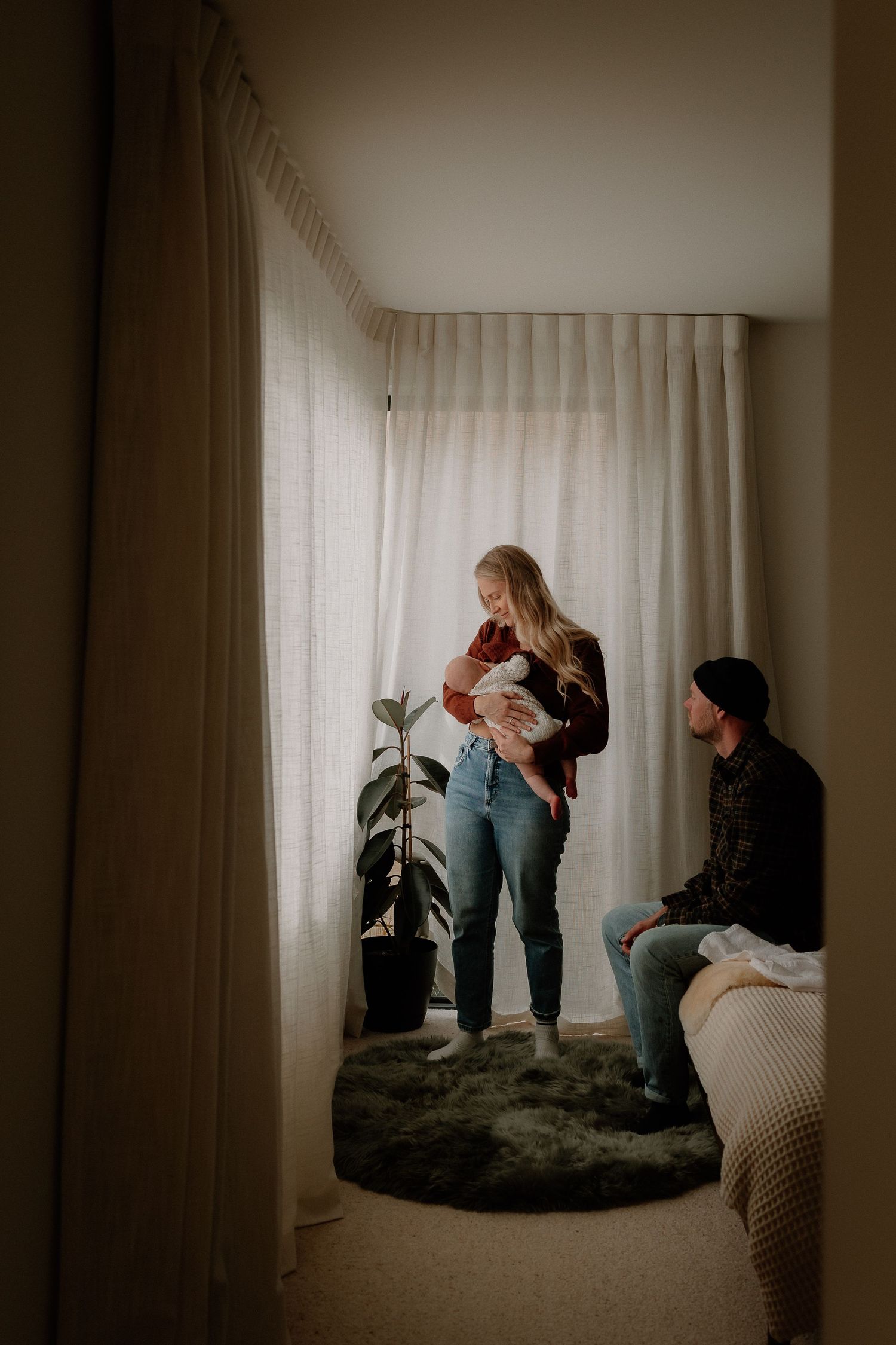 A couple shares a tender moment with their newborn baby in a softly lit bedroom with sheer curtains.