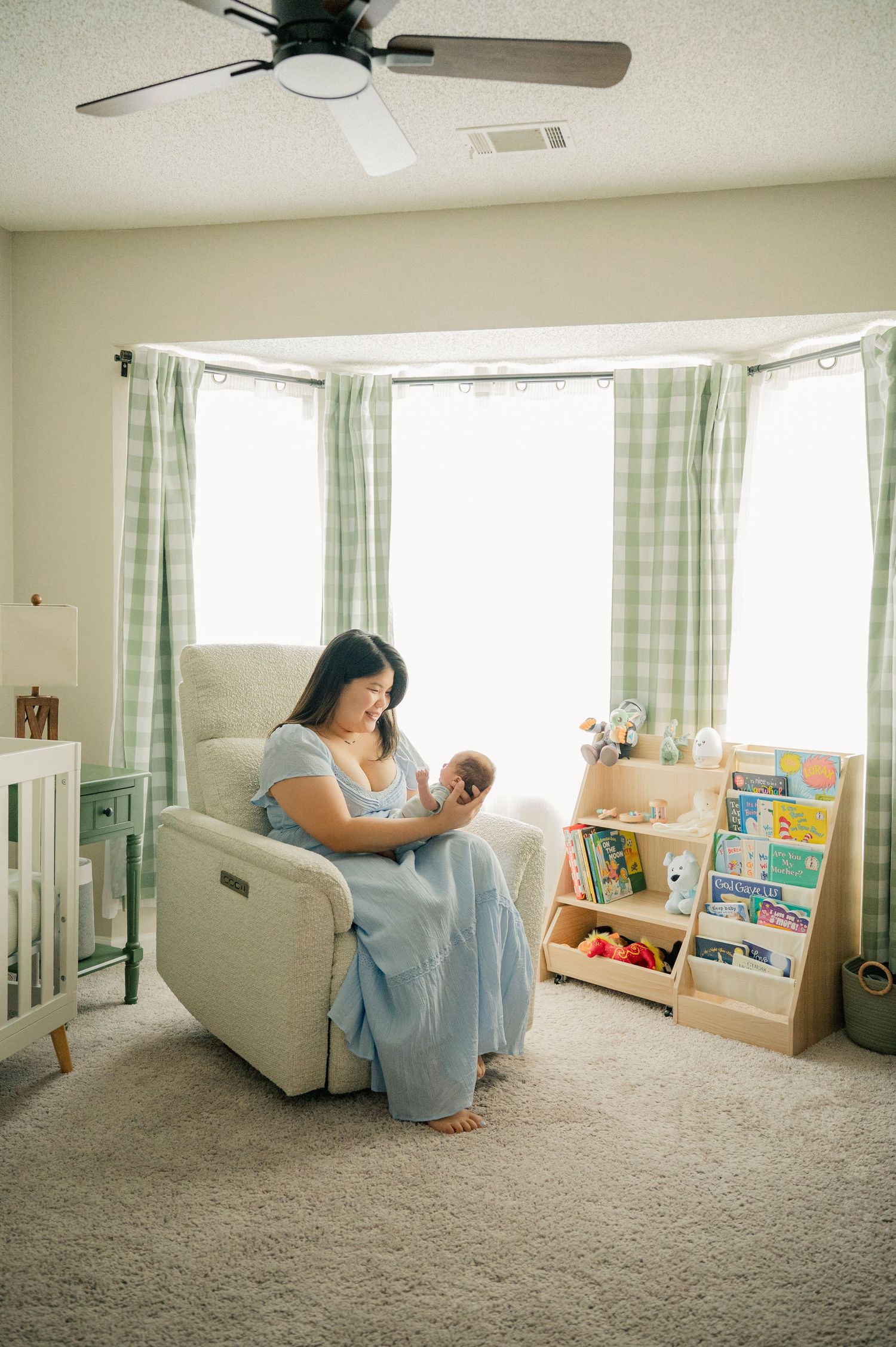 Mother sitting in nursery glider chair holding newborn baby, surrounded by bookshelf and crib with green plaid curtains.