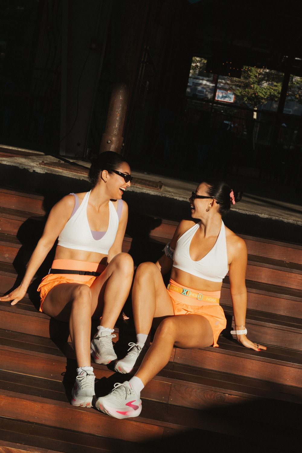 Two women in matching white sports bras and orange shorts sitting on wooden steps in sunlight.