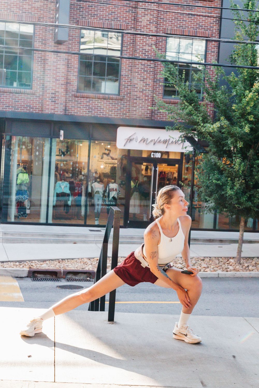 Woman in white tank and red shorts doing a lunge stretch on a city sidewalk outside a retail store.