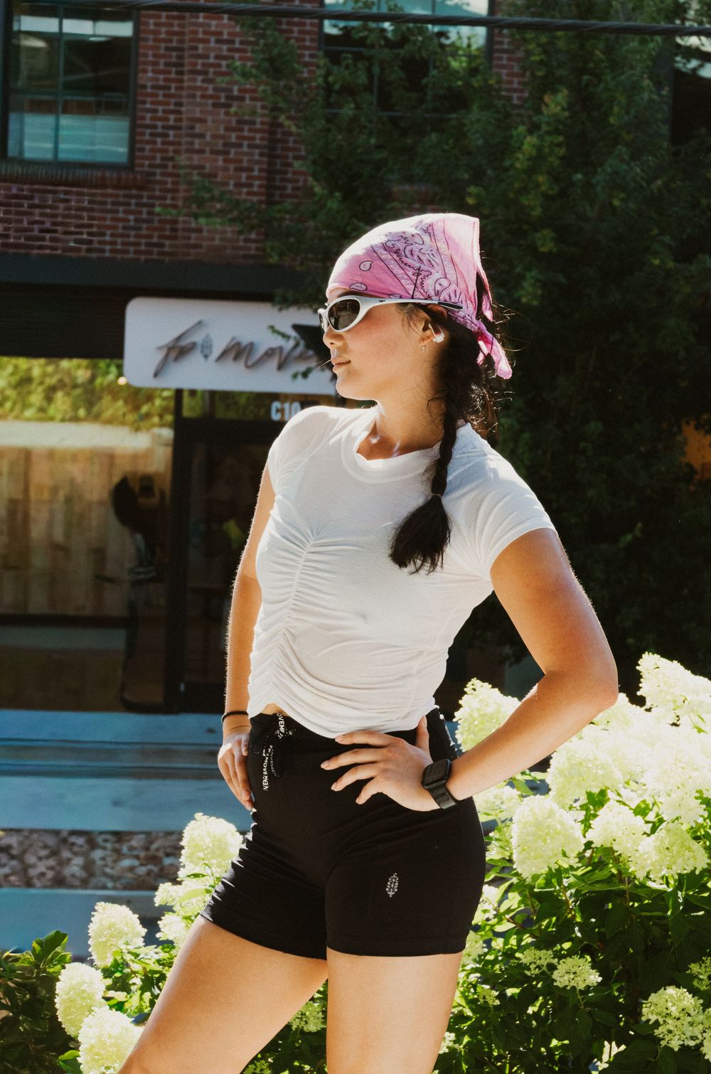 Woman in white tee, black shorts, pink bandana, and white sunglasses posing outdoors near white hydrangeas.