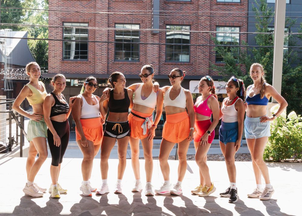 Group of nine women in colorful athletic wear posing together outdoors in front of a brick building.