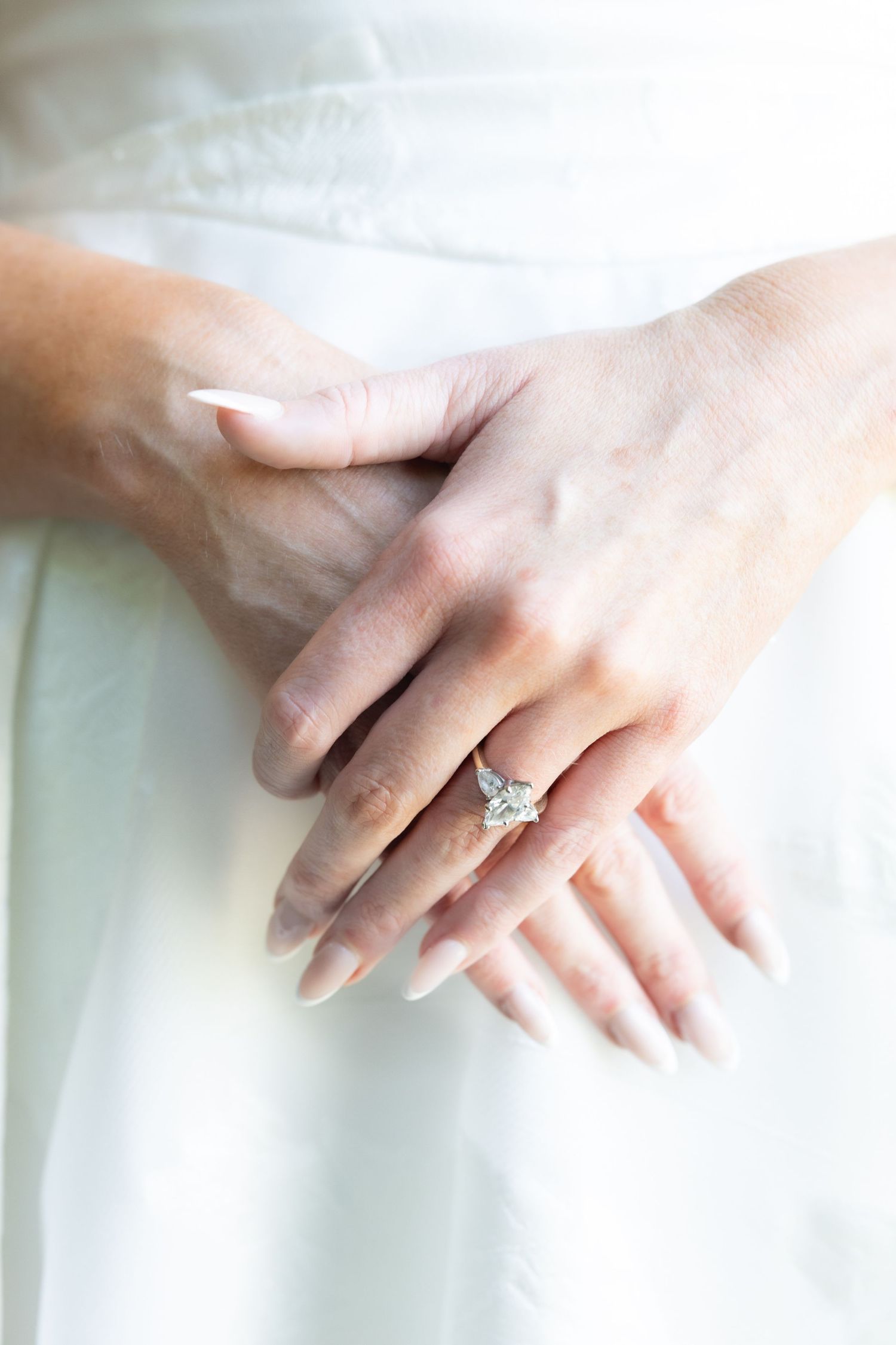 Close-up of a bride's hands folded over her white gown, featuring a stunning princess-cut diamond ring.