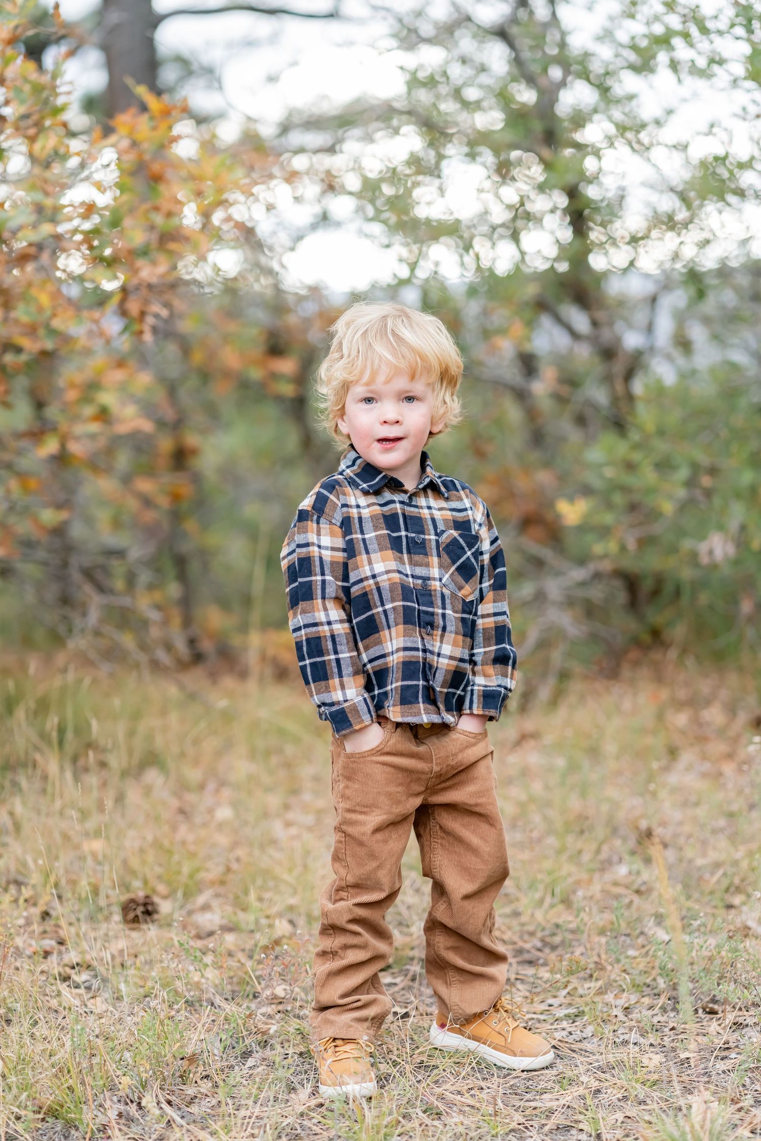 Toddler boy in plaid shirt and tan pants standing outdoors among autumn trees.