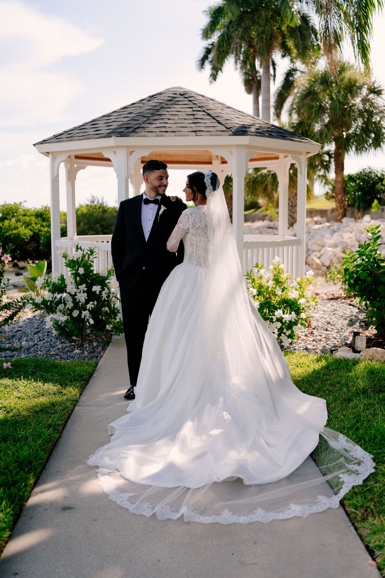 Bride in lace gown with long veil and groom in tuxedo pose by white gazebo surrounded by tropical greenery.