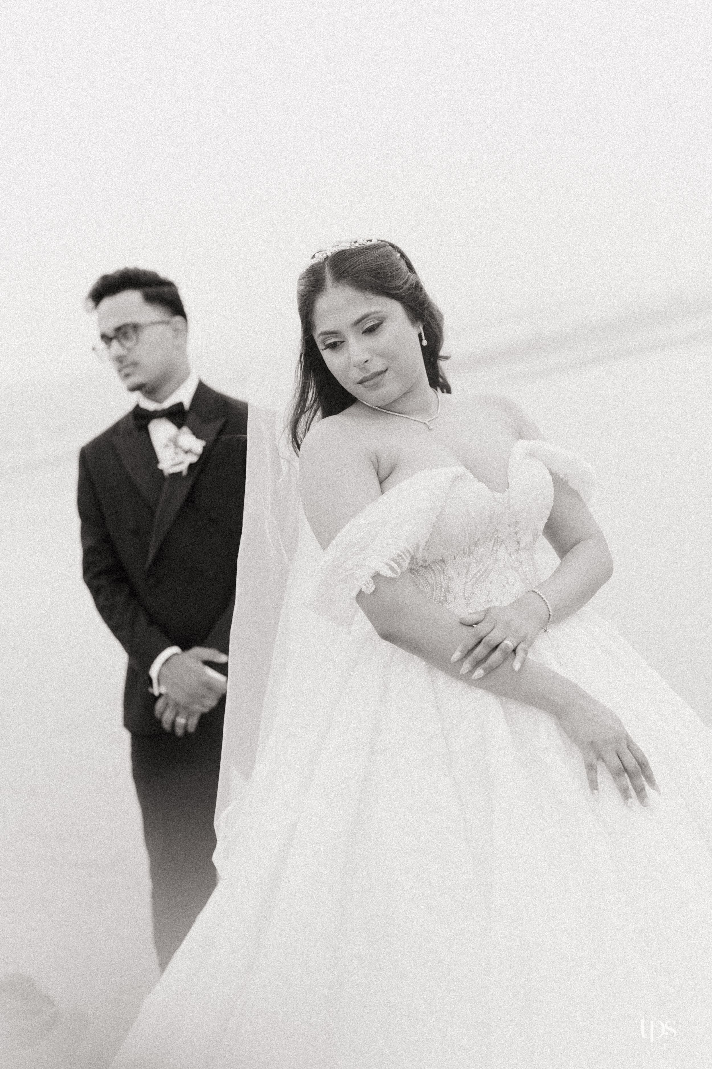 Black and white wedding photo of bride in off-shoulder gown and groom in tuxedo posing on a misty beach.