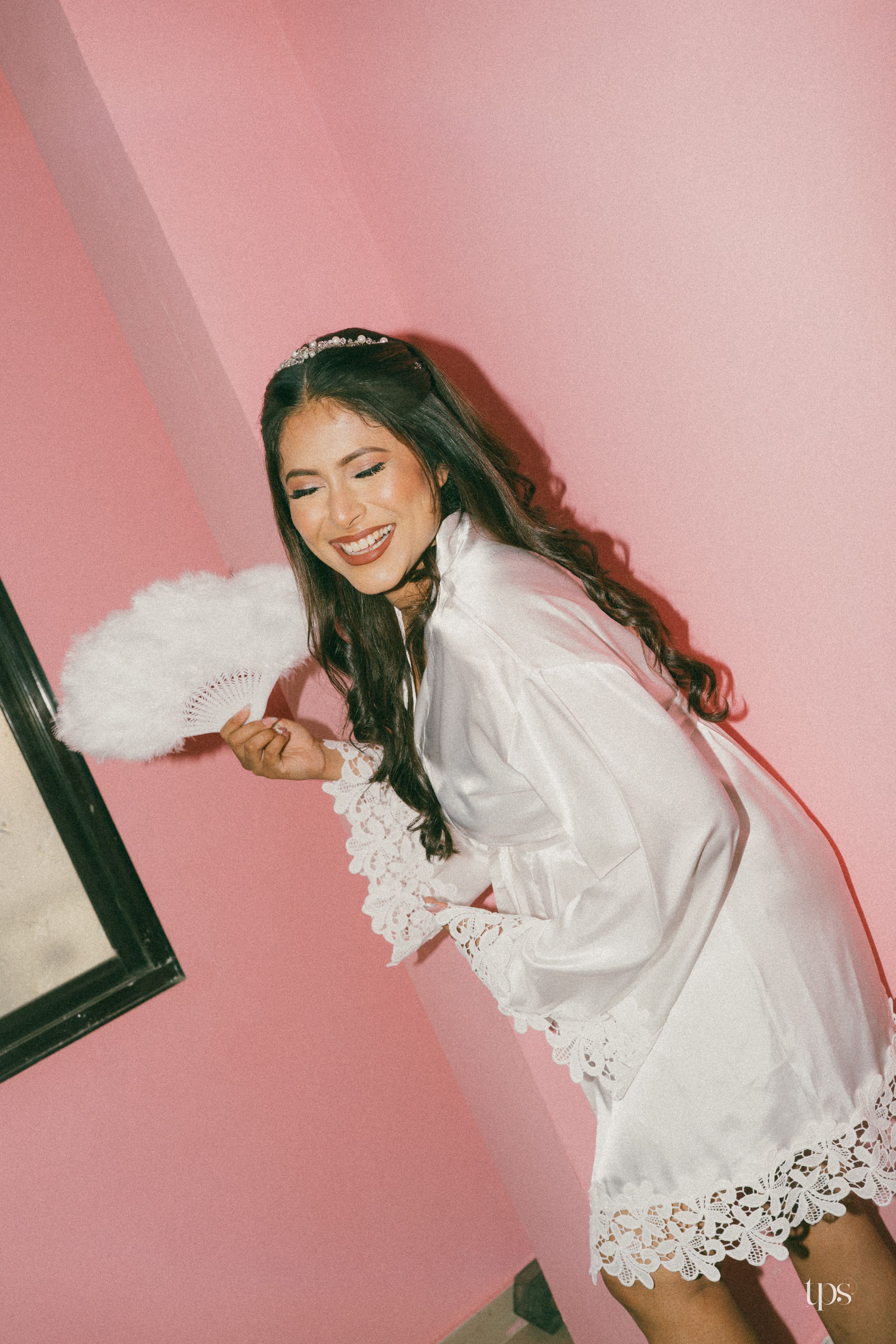 Smiling bride in white lace robe holding feather fan against pink wall with tiara.