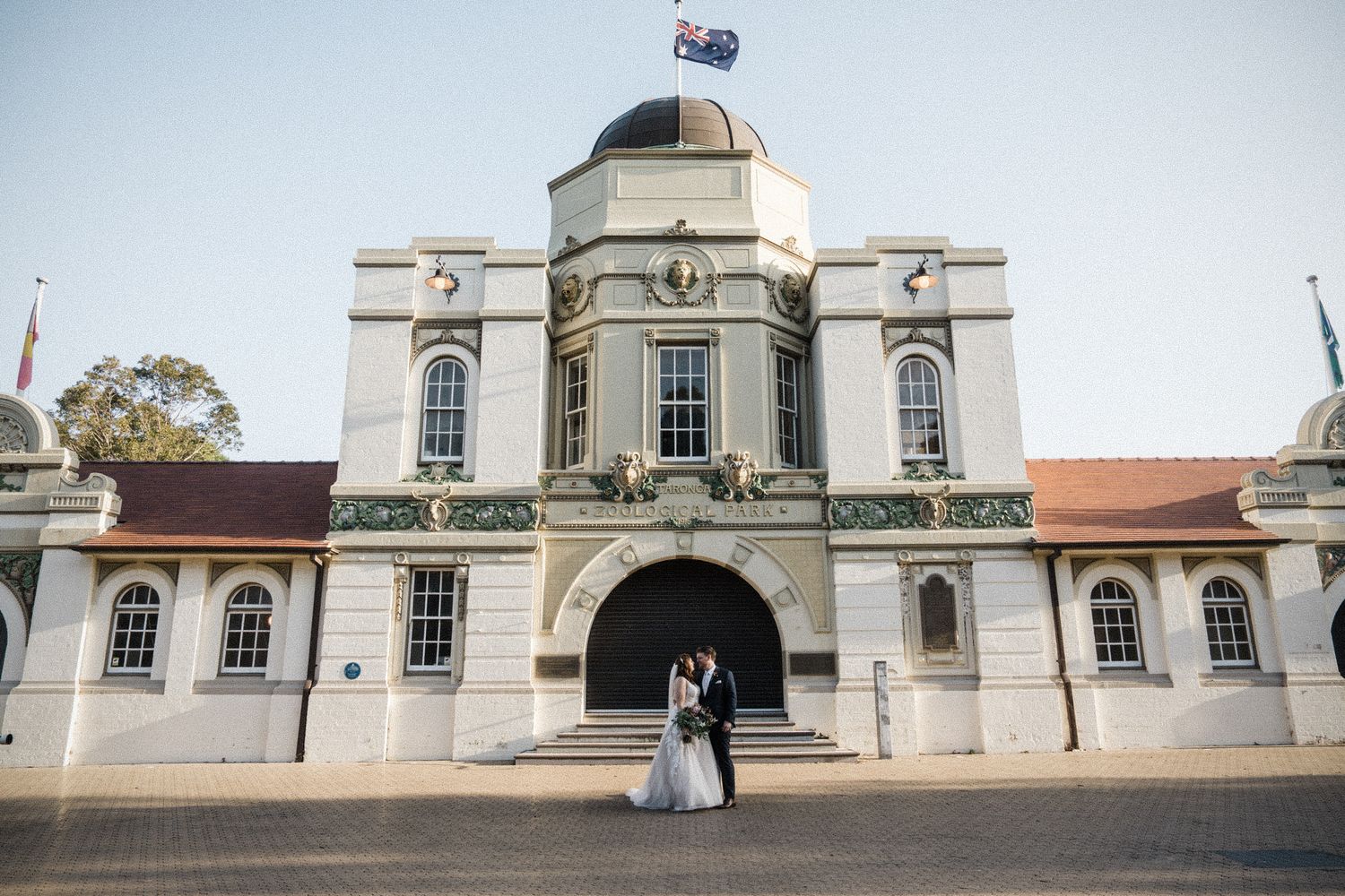 Bride and groom embracing outside an ornate white heritage building with Australian flag at their wedding.