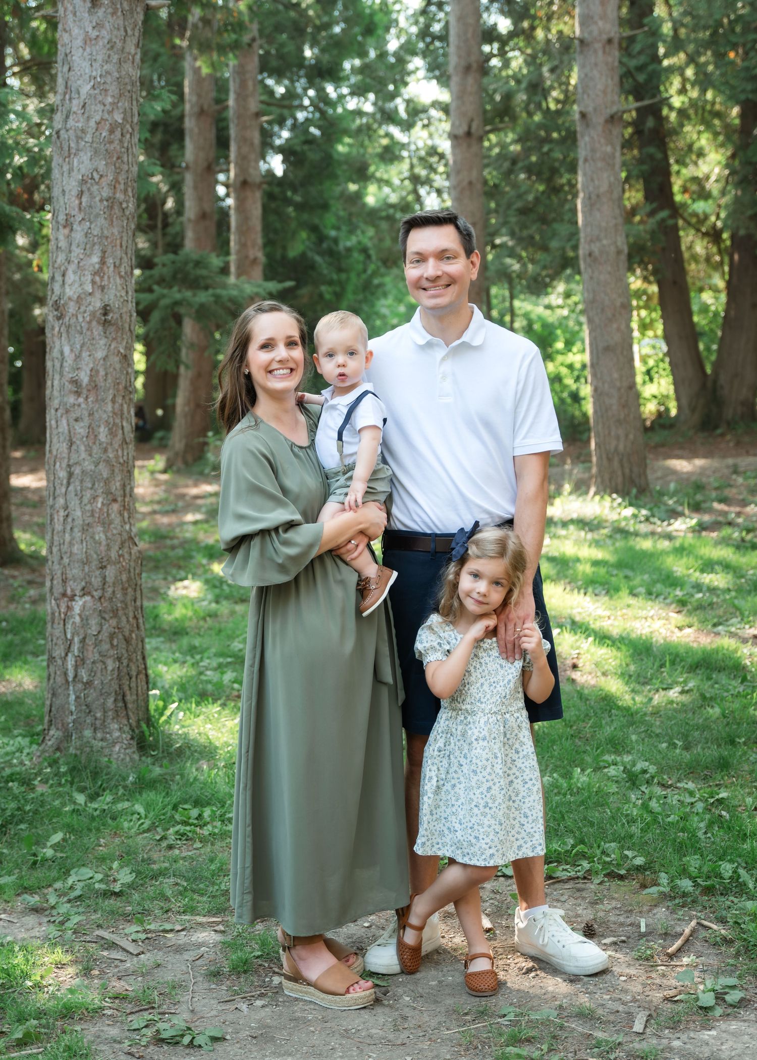 Family of four posing on a woodland path, mother in sage dress holding toddler, father in white polo, young girl in floral dress.