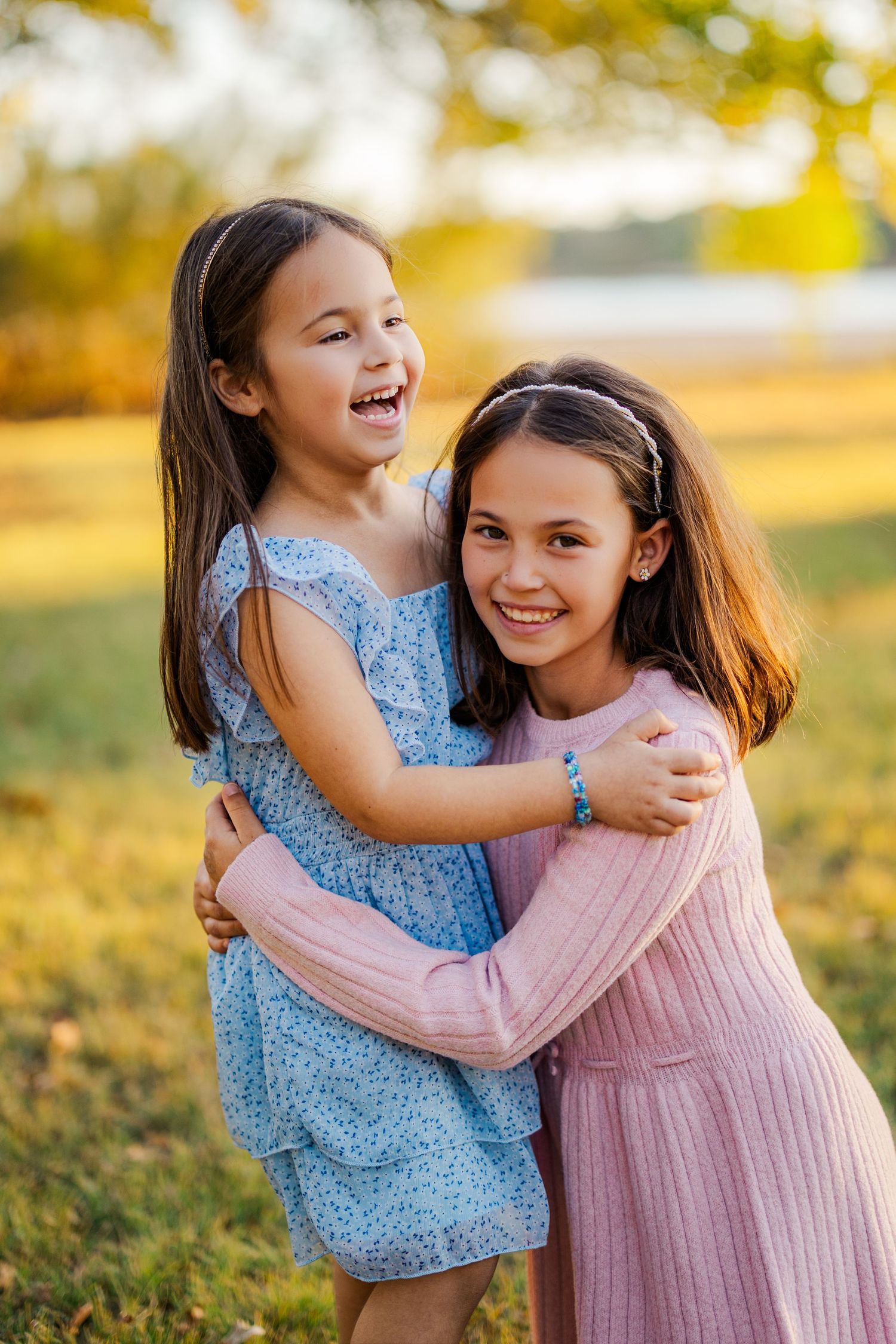 Two smiling girls in dresses hugging outdoors in a sunny park during golden hour.
