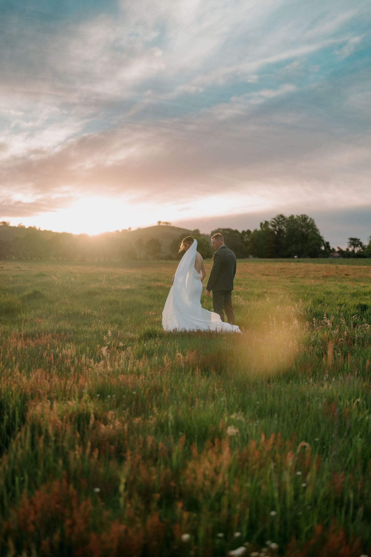Bride and groom standing in a sunlit green meadow at golden hour sunset with rolling hills in the background. The Hayshed Orange