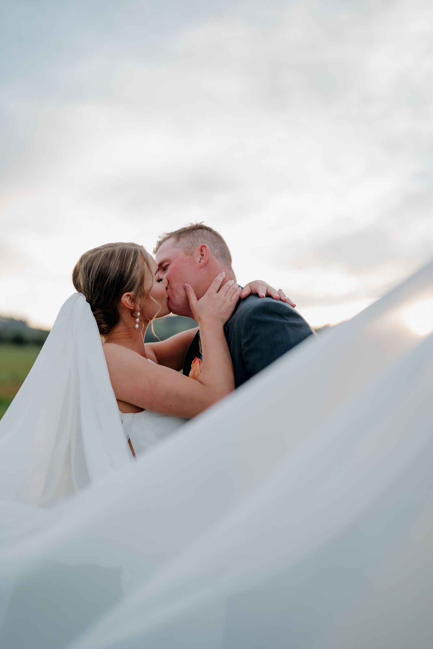 A bride and groom share a romantic kiss outdoors, framed by a flowing white veil against a cloudy sky. The Hayshed Orange