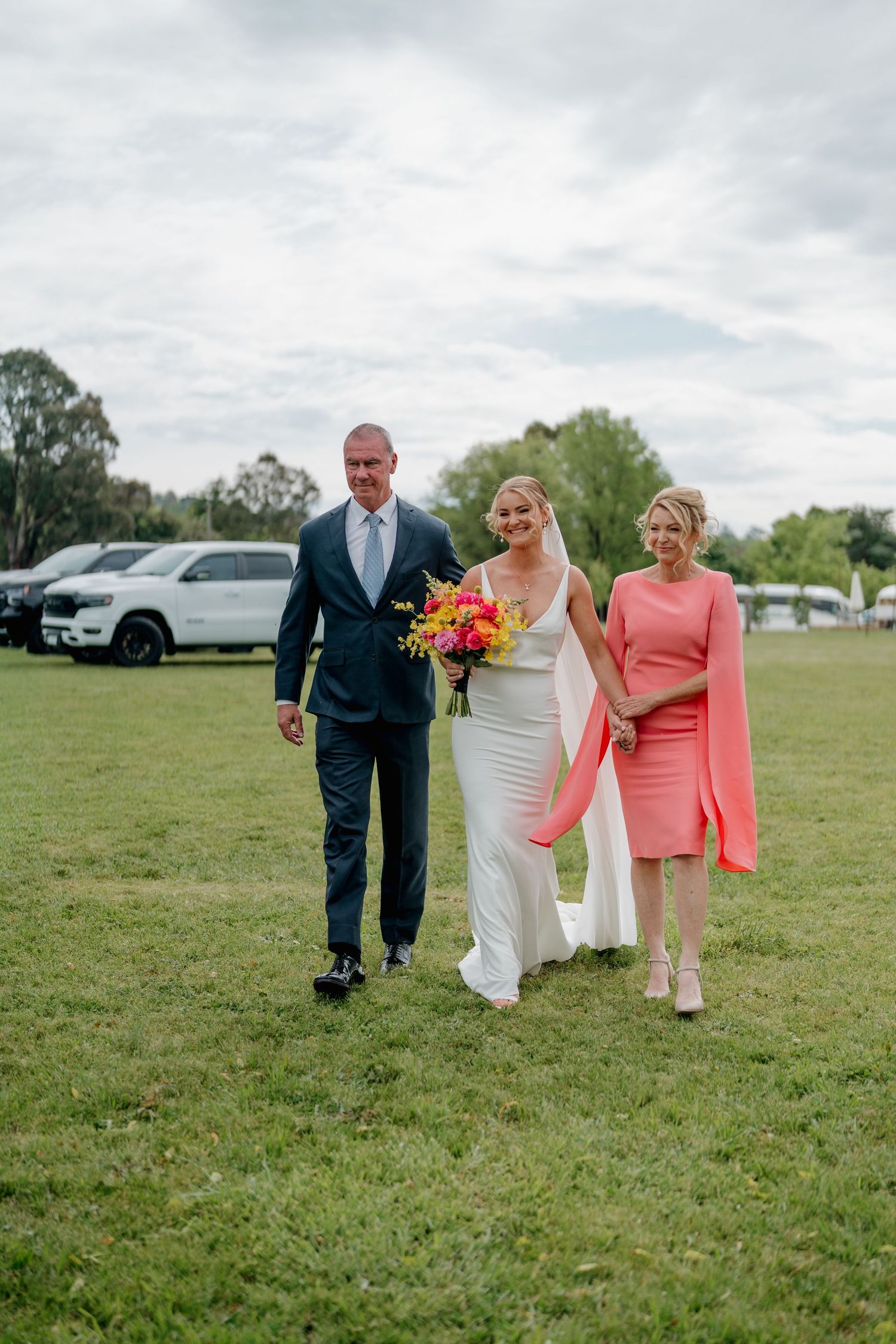 Bride in white gown walks with parents across green lawn, holding colorful bouquet, father in navy suit, mother in coral dress. The Hayshed