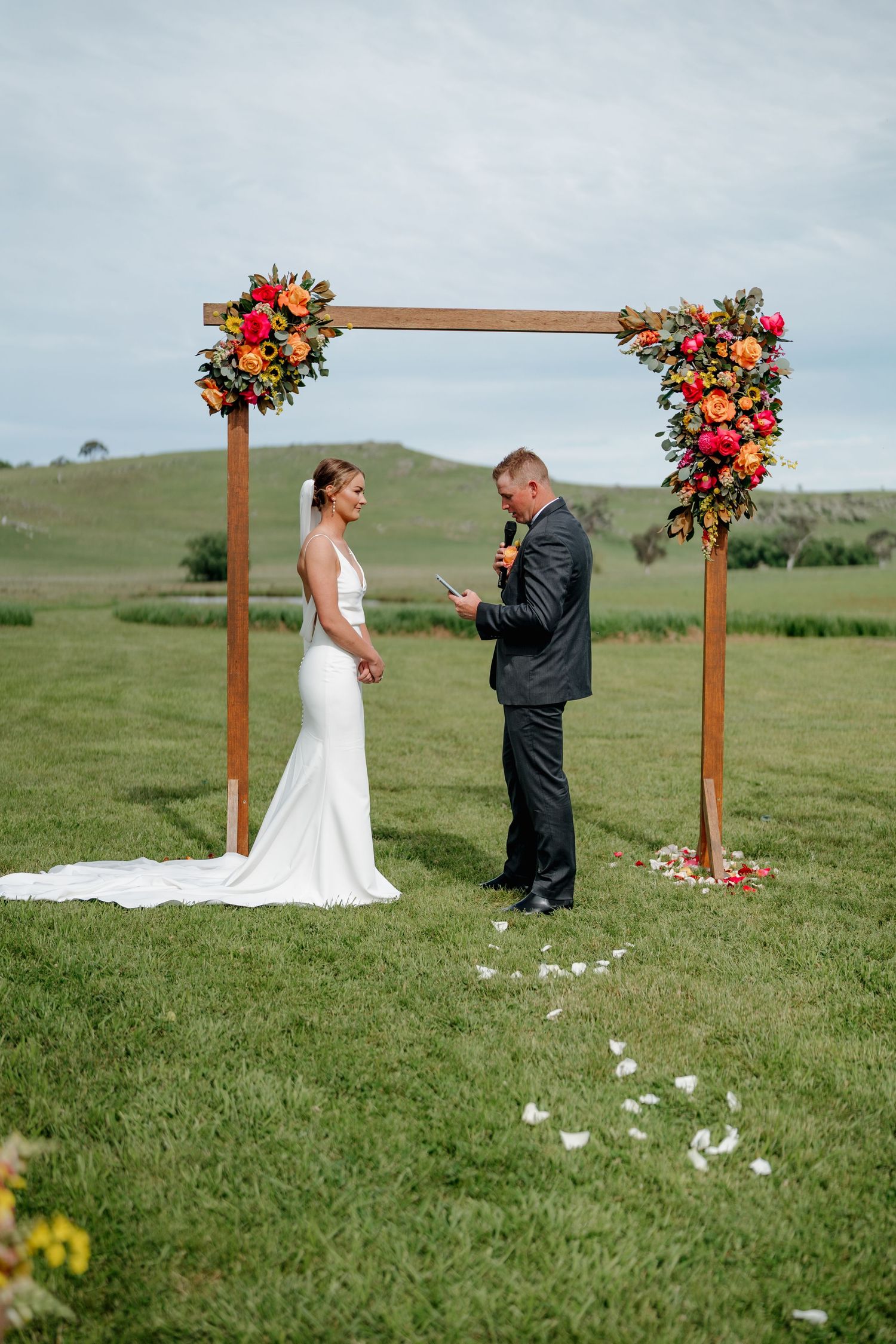Bride and groom exchange vows under a wooden arch adorned with colorful flowers at an outdoor wedding ceremony. The Hayshed Orange