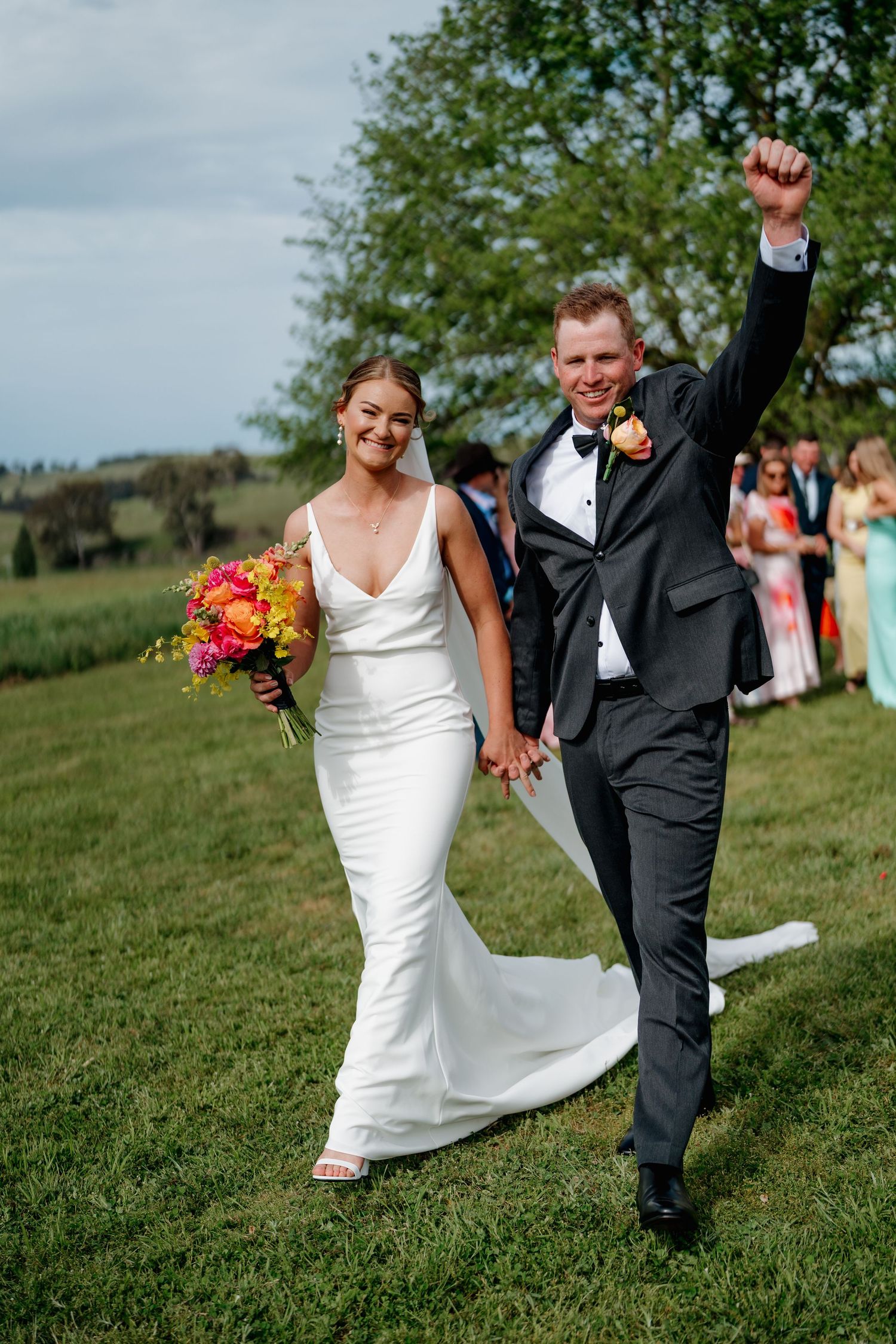 Joyful newlyweds walk hand-in-hand on green lawn, bride in white gown with colorful bouquet, groom in black suit raising fist. The Hayshed