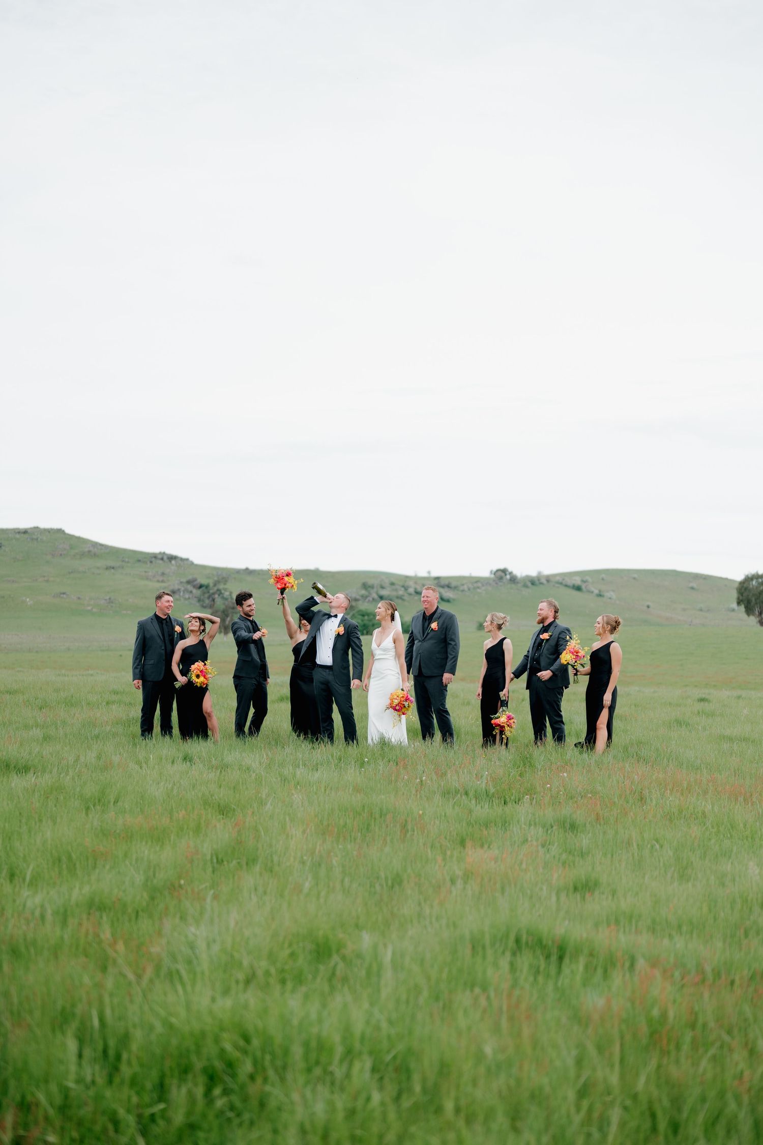 Wedding party in black attire with bride in white gown, holding orange bouquets in a lush green meadow. The Hayshed Orange