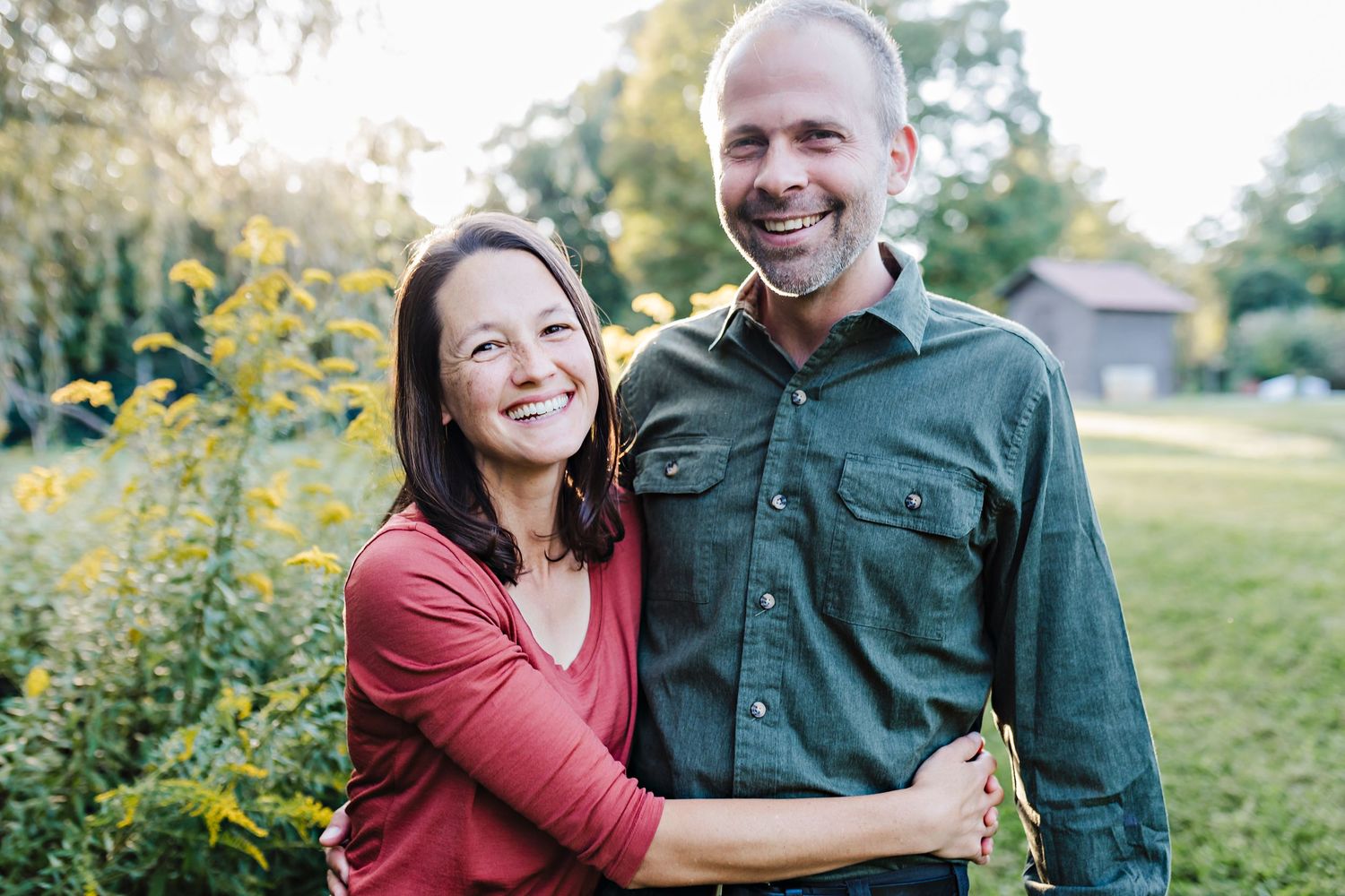 Smiling couple embracing outdoors in a lush garden with yellow wildflowers and trees in the background.