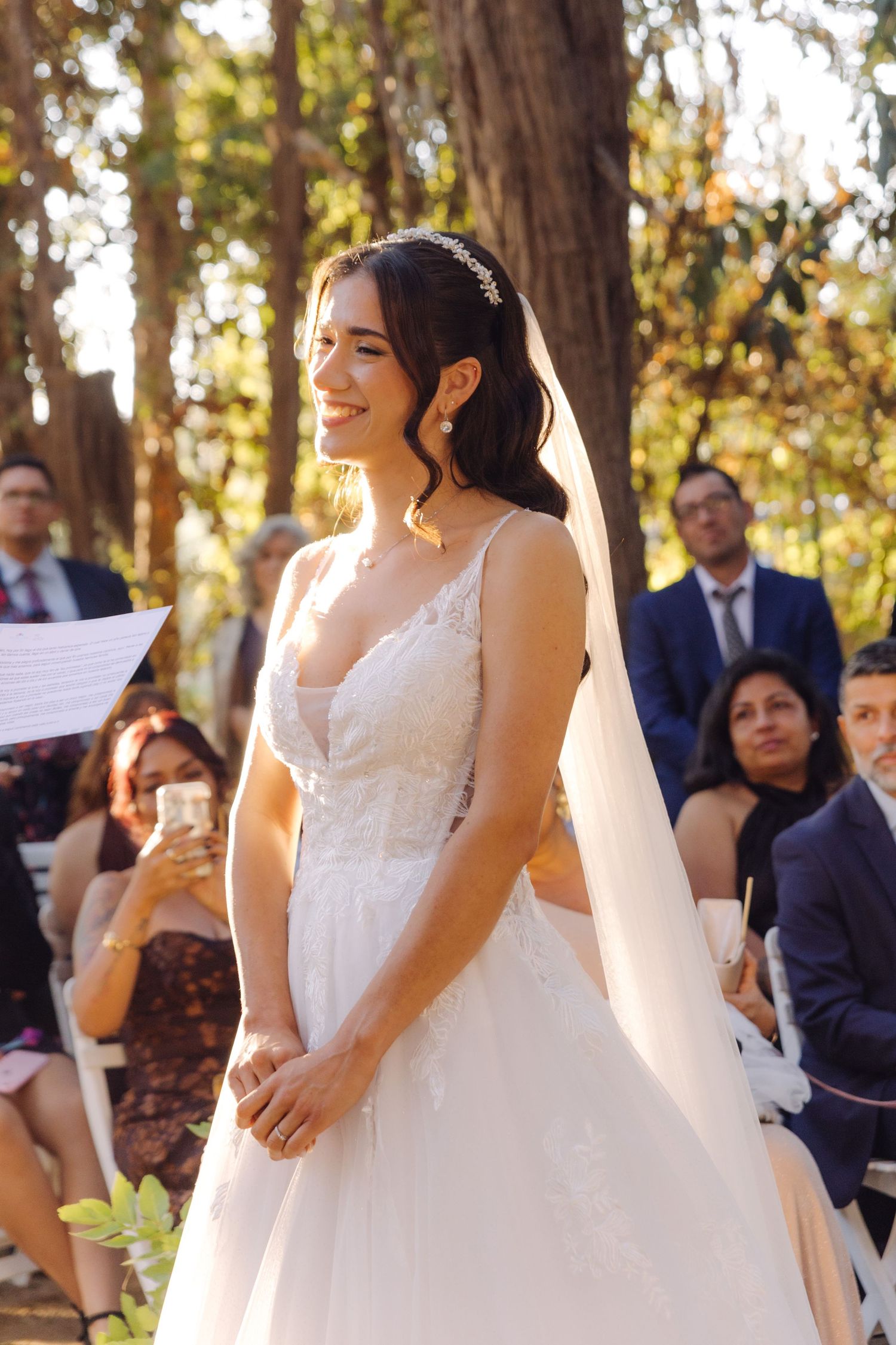 Beautiful bride in elegant white wedding dress smiling during outdoor forest ceremony.