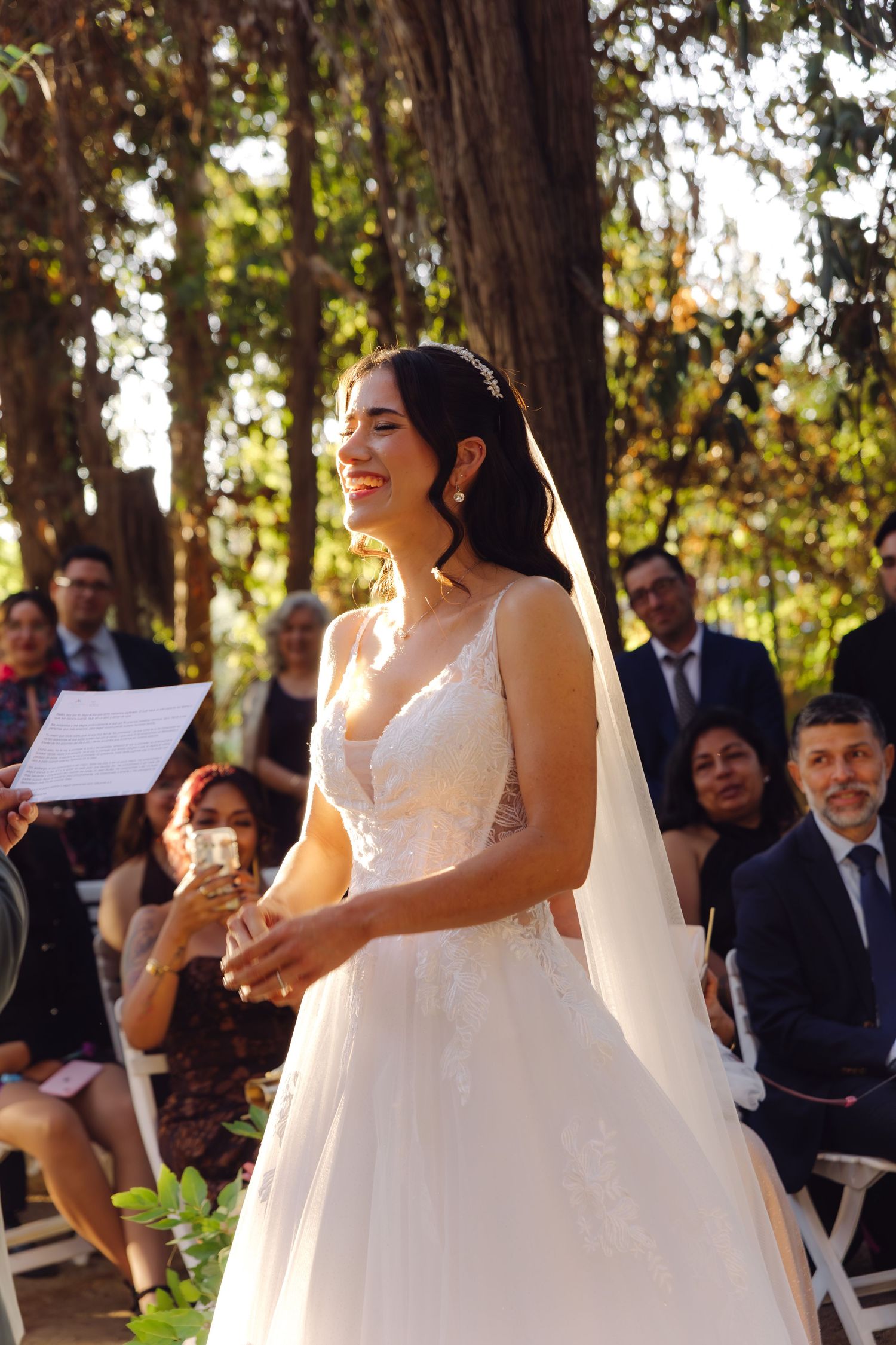 Radiant bride in white lace gown laughing during sunlit outdoor wedding ceremony.