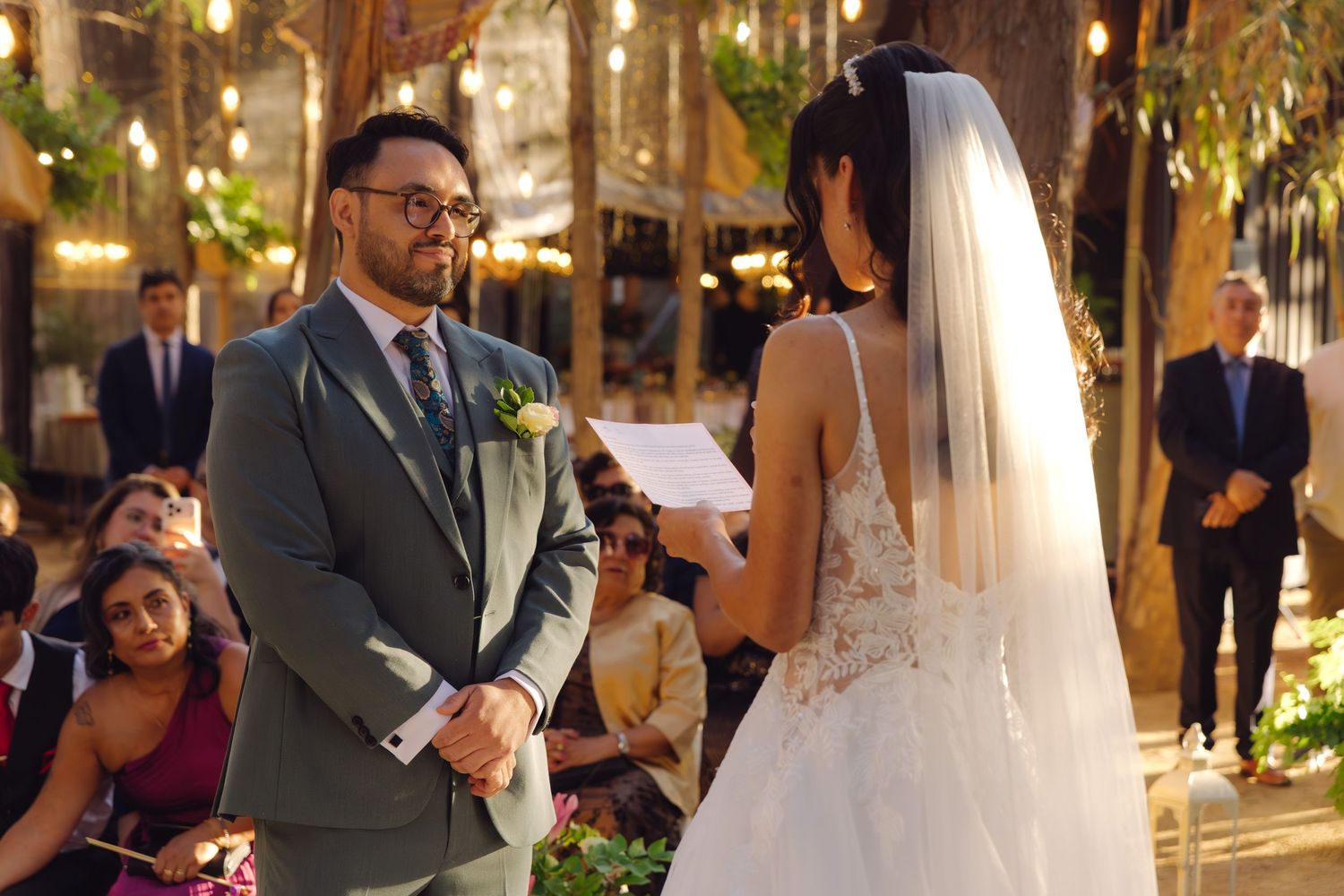 Groom in gray suit listening attentively as bride recites vows at outdoor wedding.