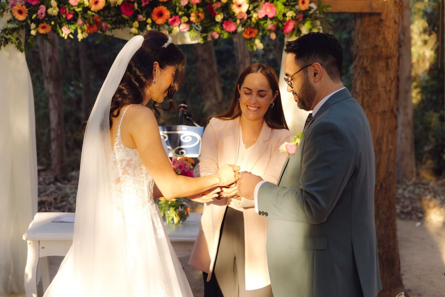 Bride placing wedding ring on grooms finger under floral arch with officiant.