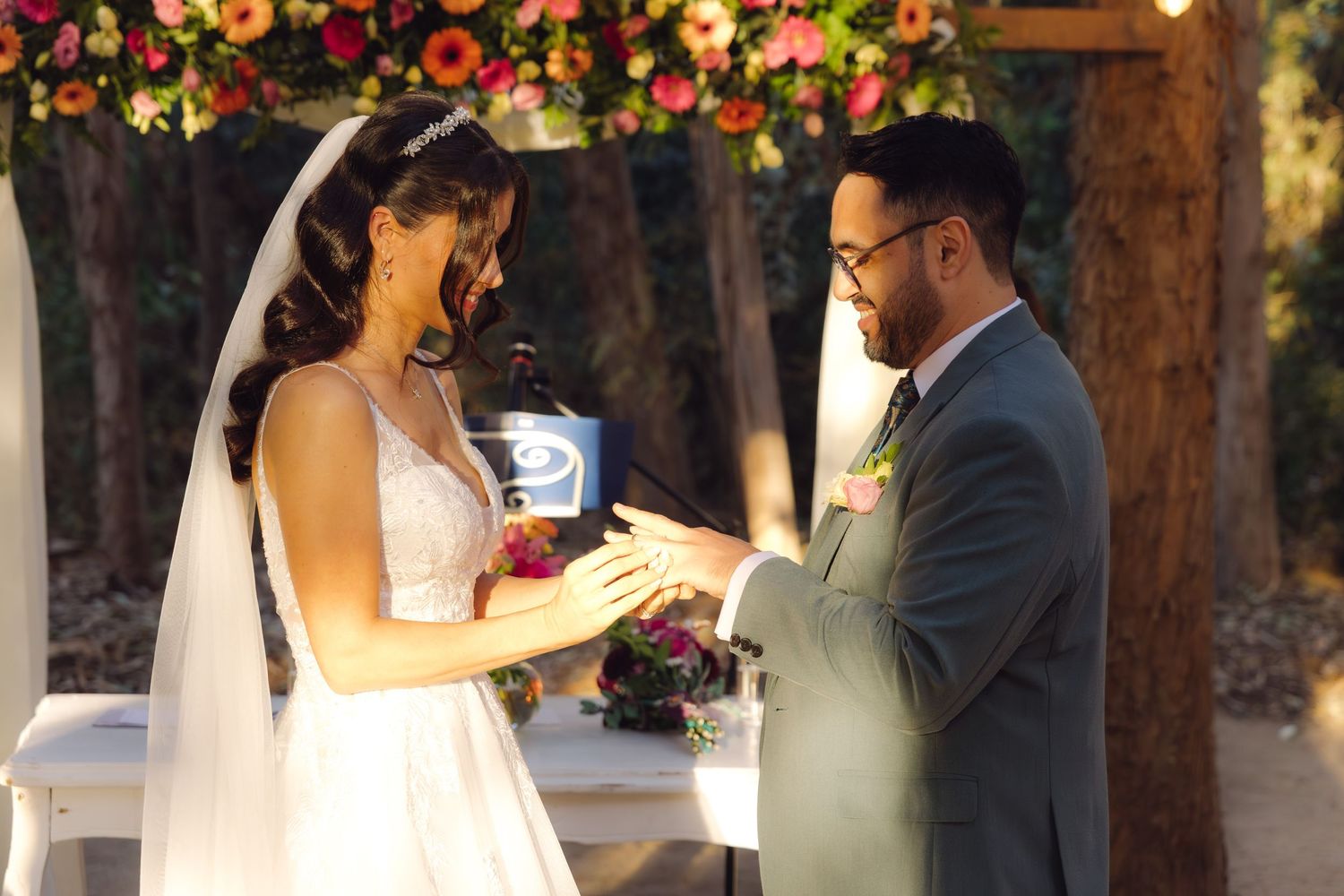 Bride and groom exchanging rings under colorful floral arch at outdoor wedding.