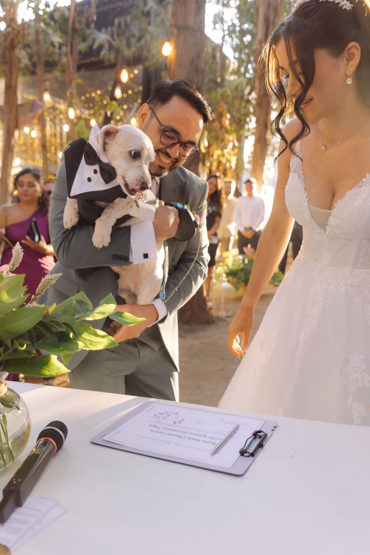 Groom holding small dog in tuxedo outfit while bride stands at signing table at outdoor wedding.